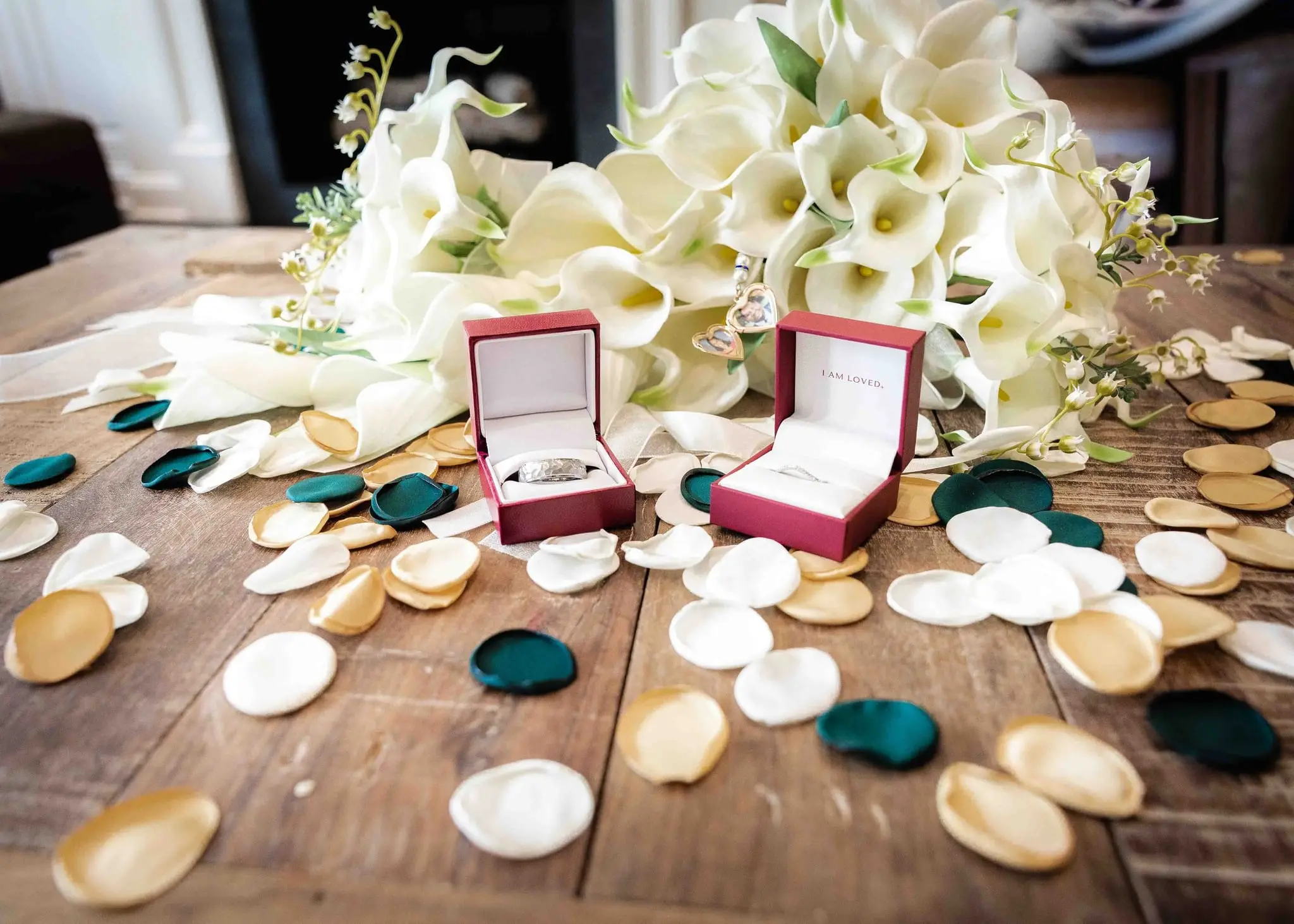 Ring boxes on a rustic table beside a calla lily bouquet and scattered green, gold and white petals