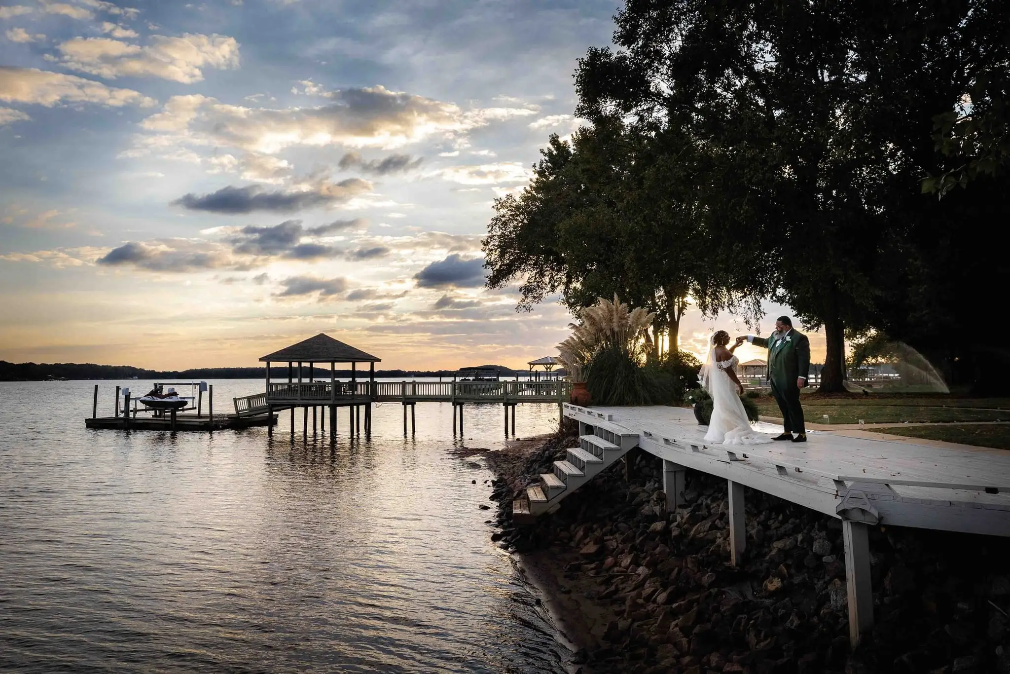 Couple standing together on a white lakeside pier beneath a pastel sunset sky