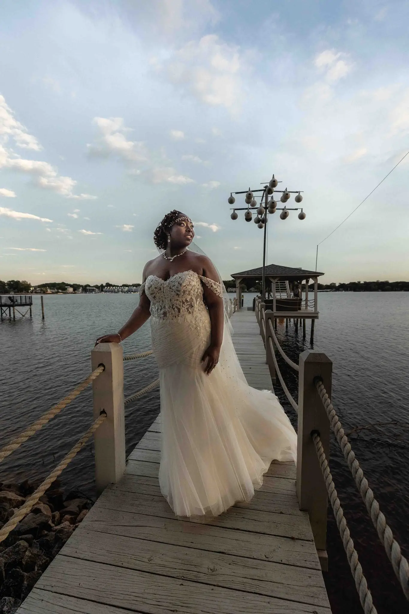 Bride in a fitted lace gown standing on a rope-railed dock over the lake at Whispering Oaks
