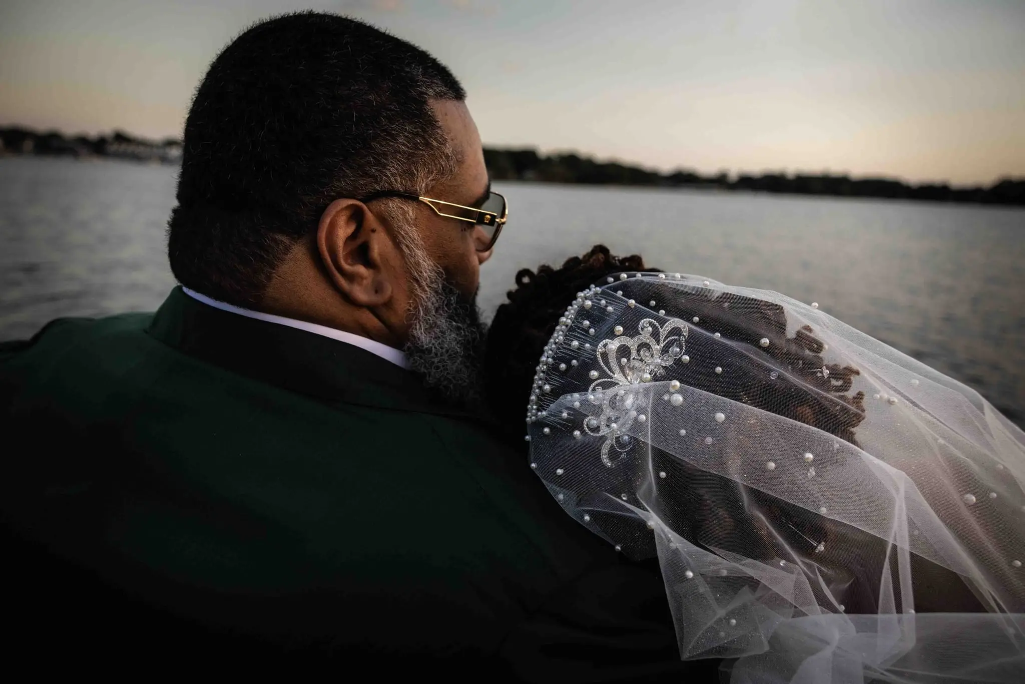 Groom in a green suit leaning close to the bride, her pearl-studded veil blowing, on a boat at dusk