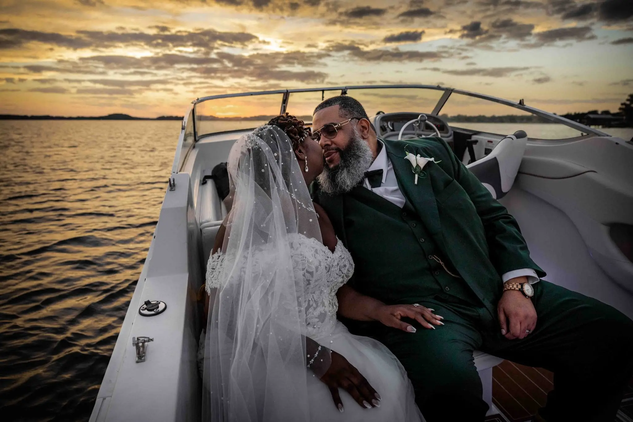 Couple leaning in for a kiss on a boat as the sun sets, her veil blowing in the breeze