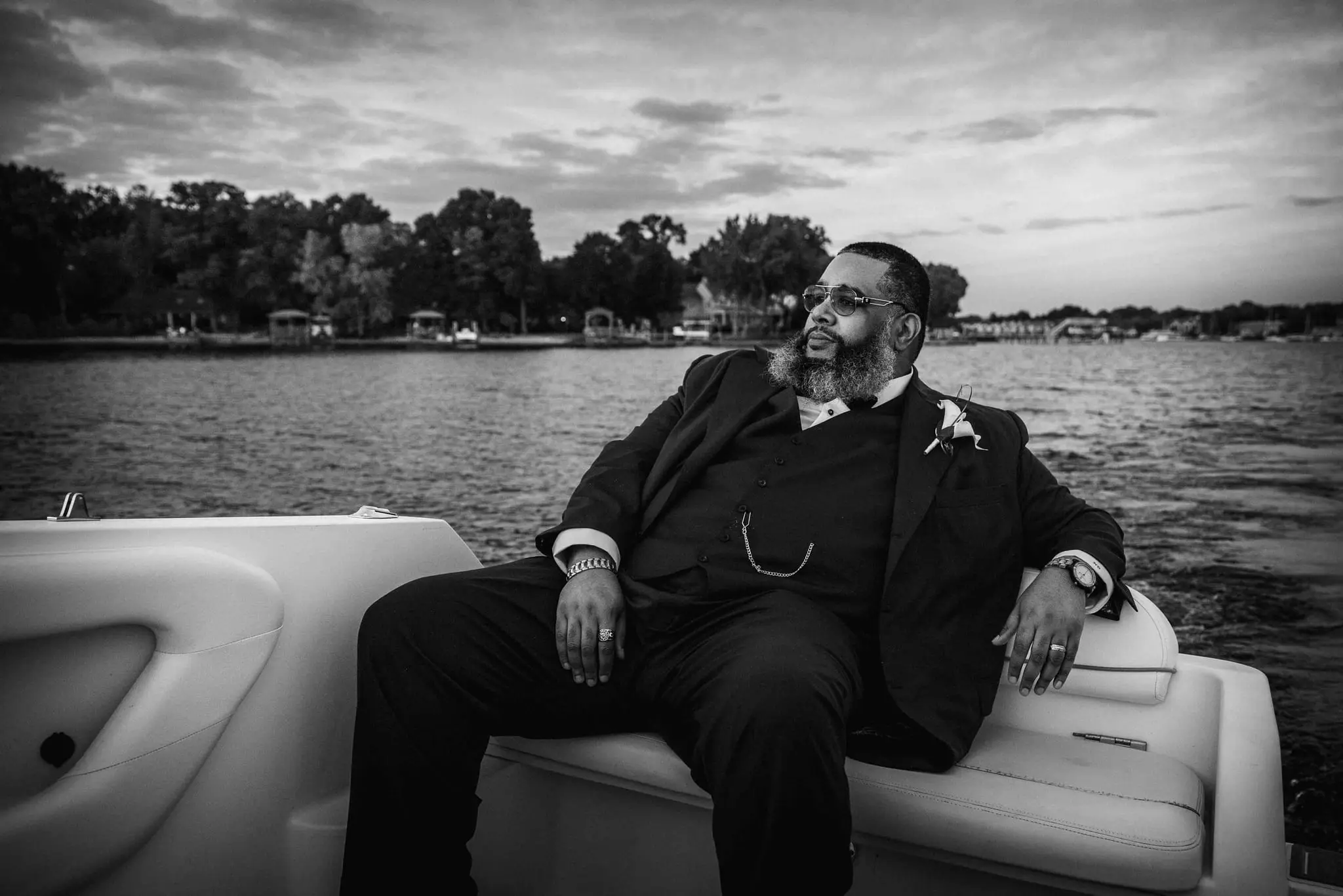 Black and white portrait of the groom in a vest, reclined on the boat seat gazing across the lake