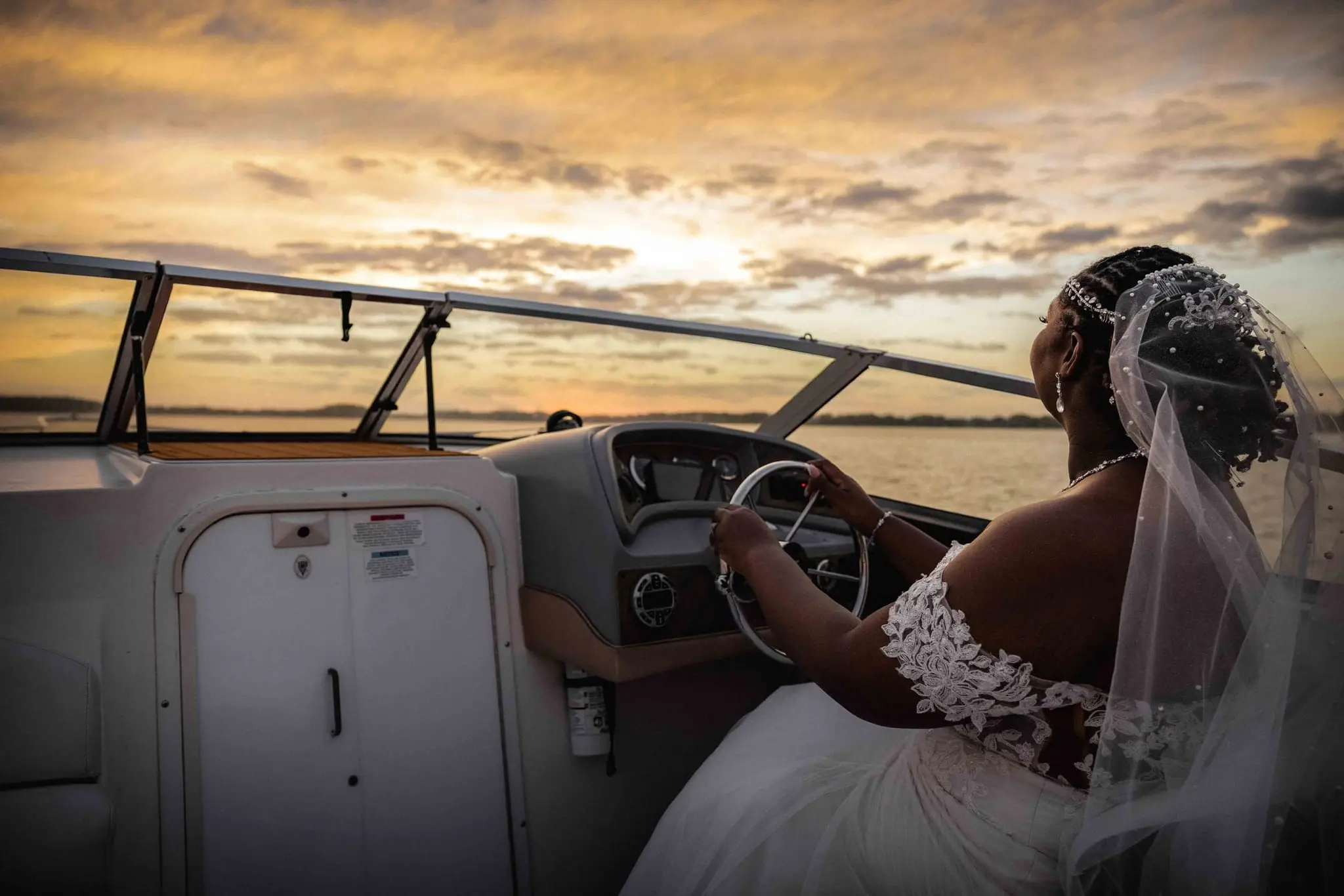 Bride at the boat's steering wheel looking toward the sunset, veil trailing