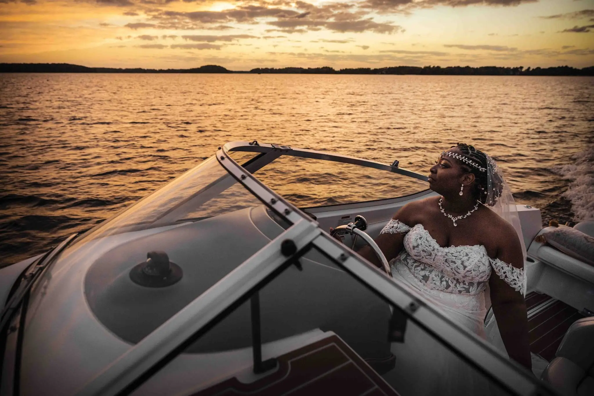 Bride in a lace gown seated at the boat's wheel, looking back over the lake in golden light