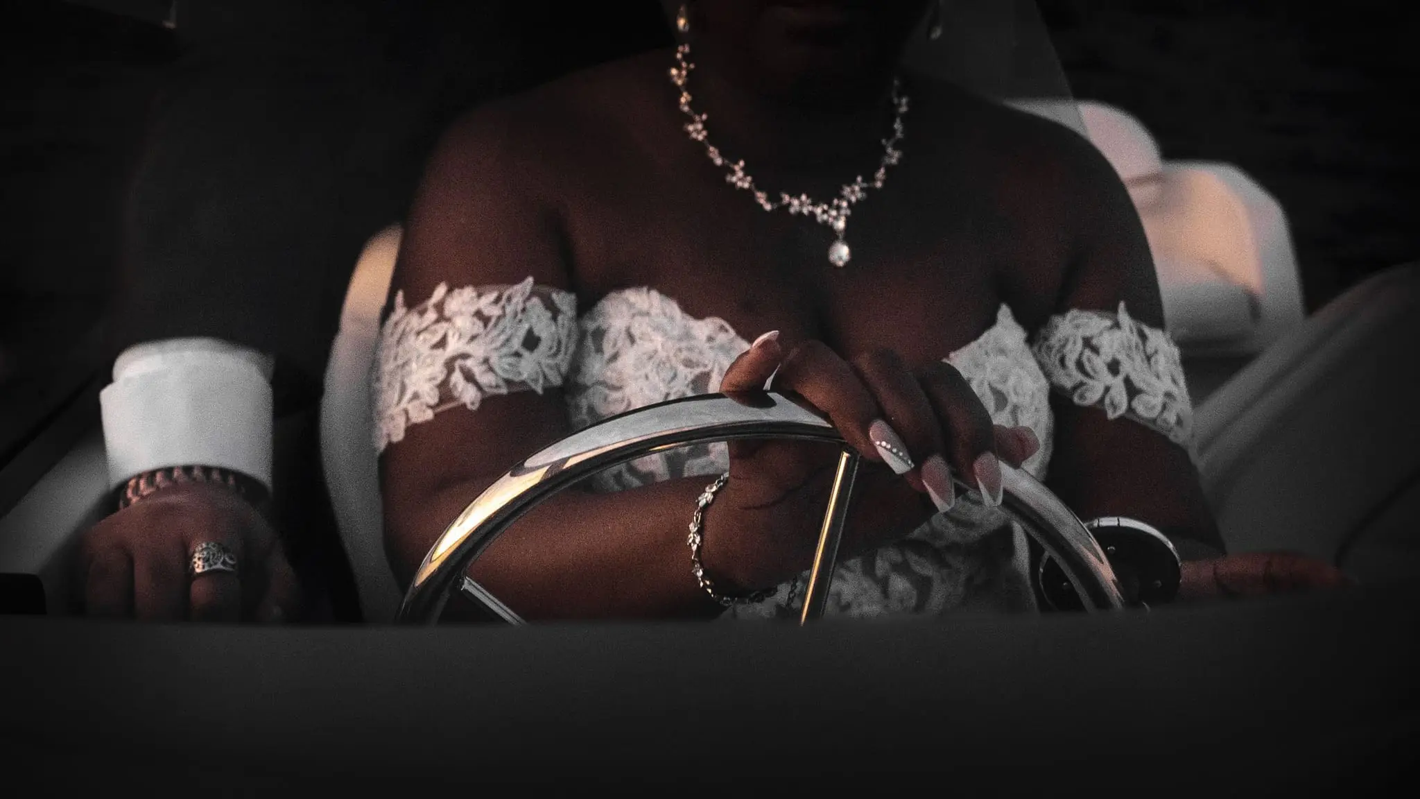 Close detail of the bride's lace sleeves, crystal necklace and hand on the steering wheel
