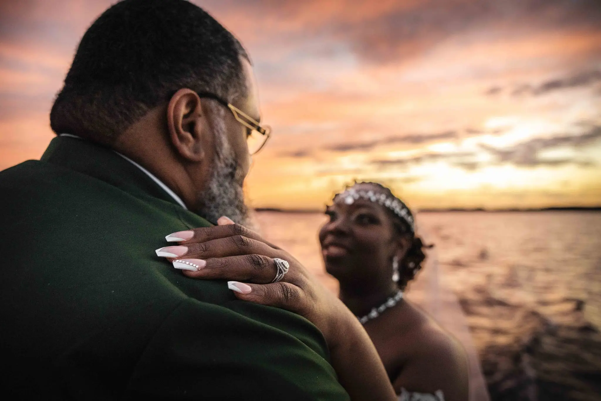 Bride's manicured hand on the groom's green jacket as she smiles at him against a pink sunset