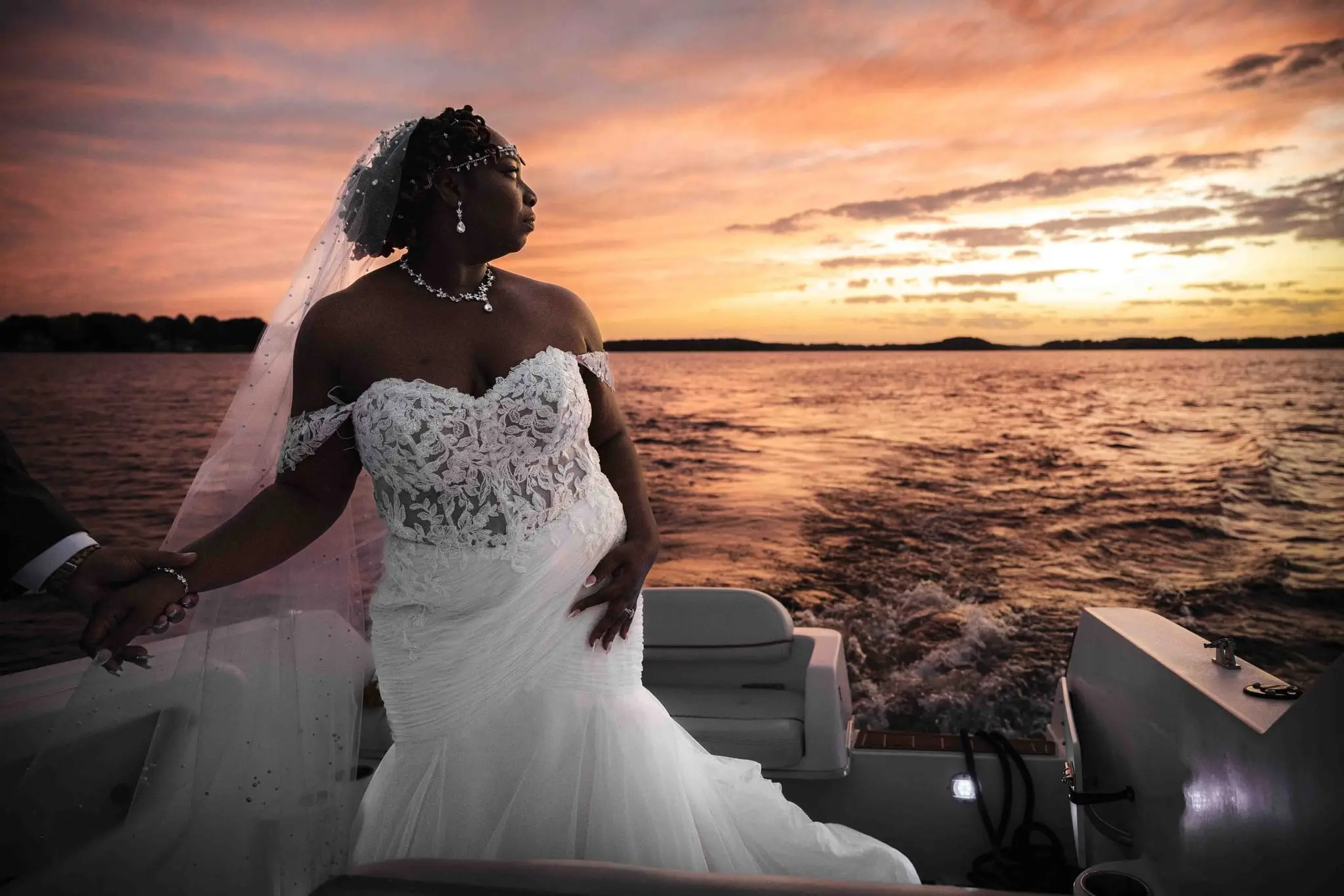 Bride standing on the boat deck in her lace gown, gazing at the lake under a pink and orange sky