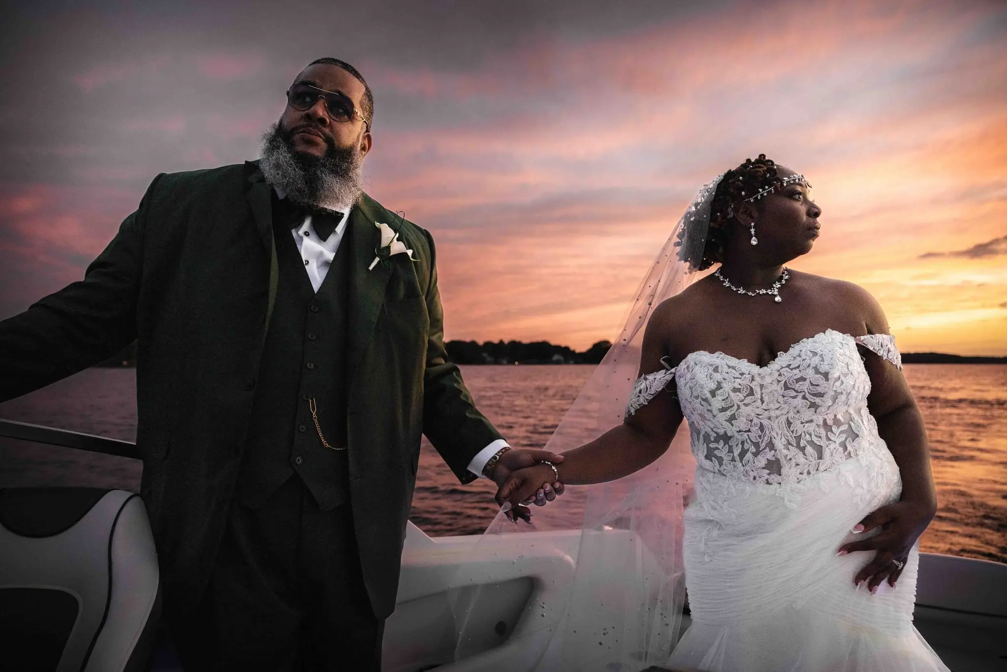 Couple holding hands on the boat, the bride's other hand on her hip, a sunset glowing behind