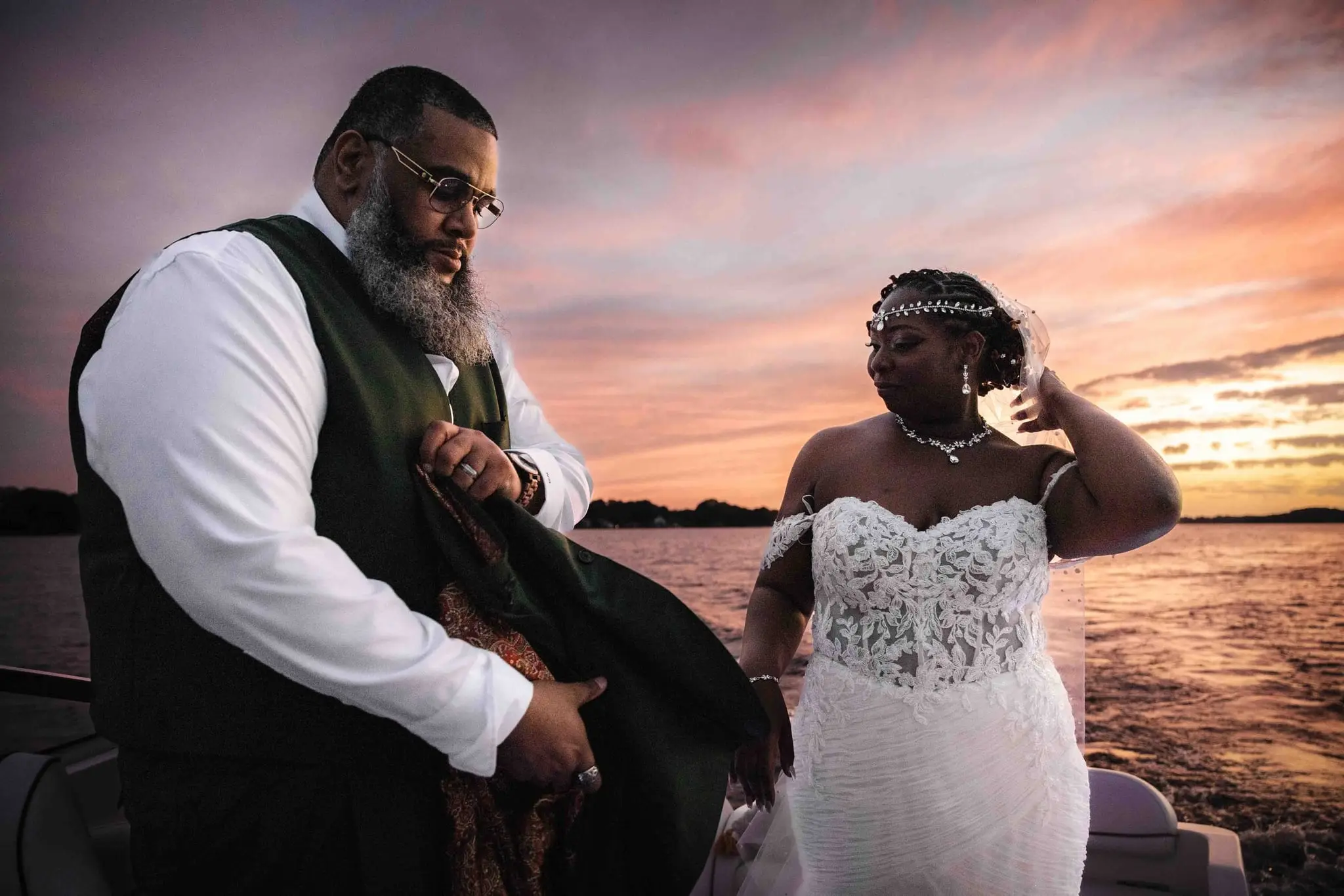 Groom holding his green jacket open to wrap the bride on the boat at sunset