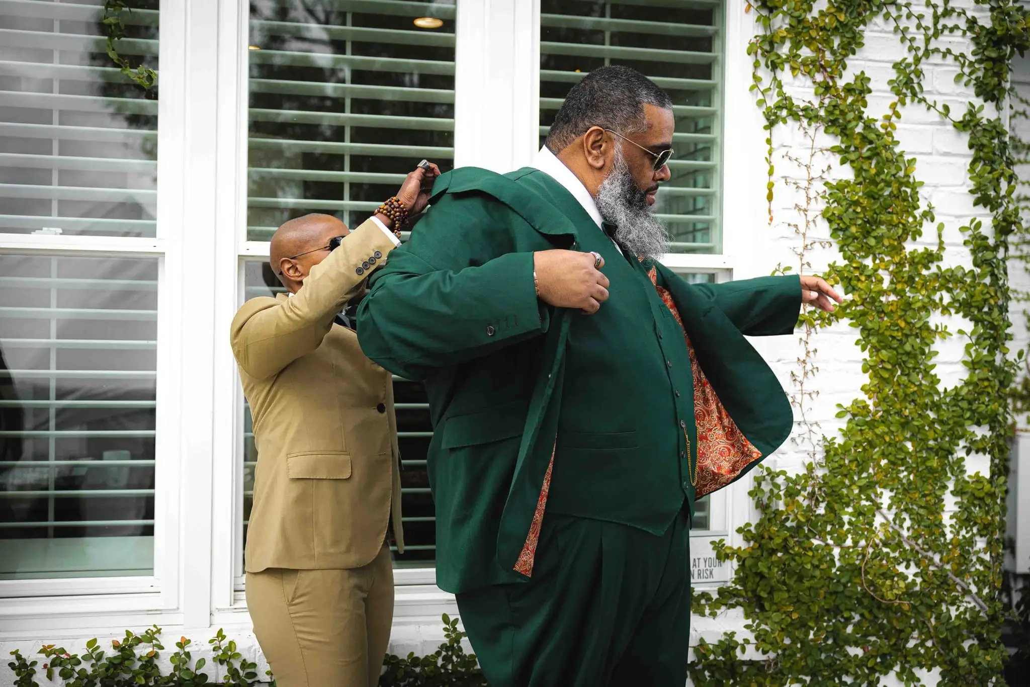 A groomsman in a tan suit helps the groom into his emerald jacket beside an ivy-covered wall
