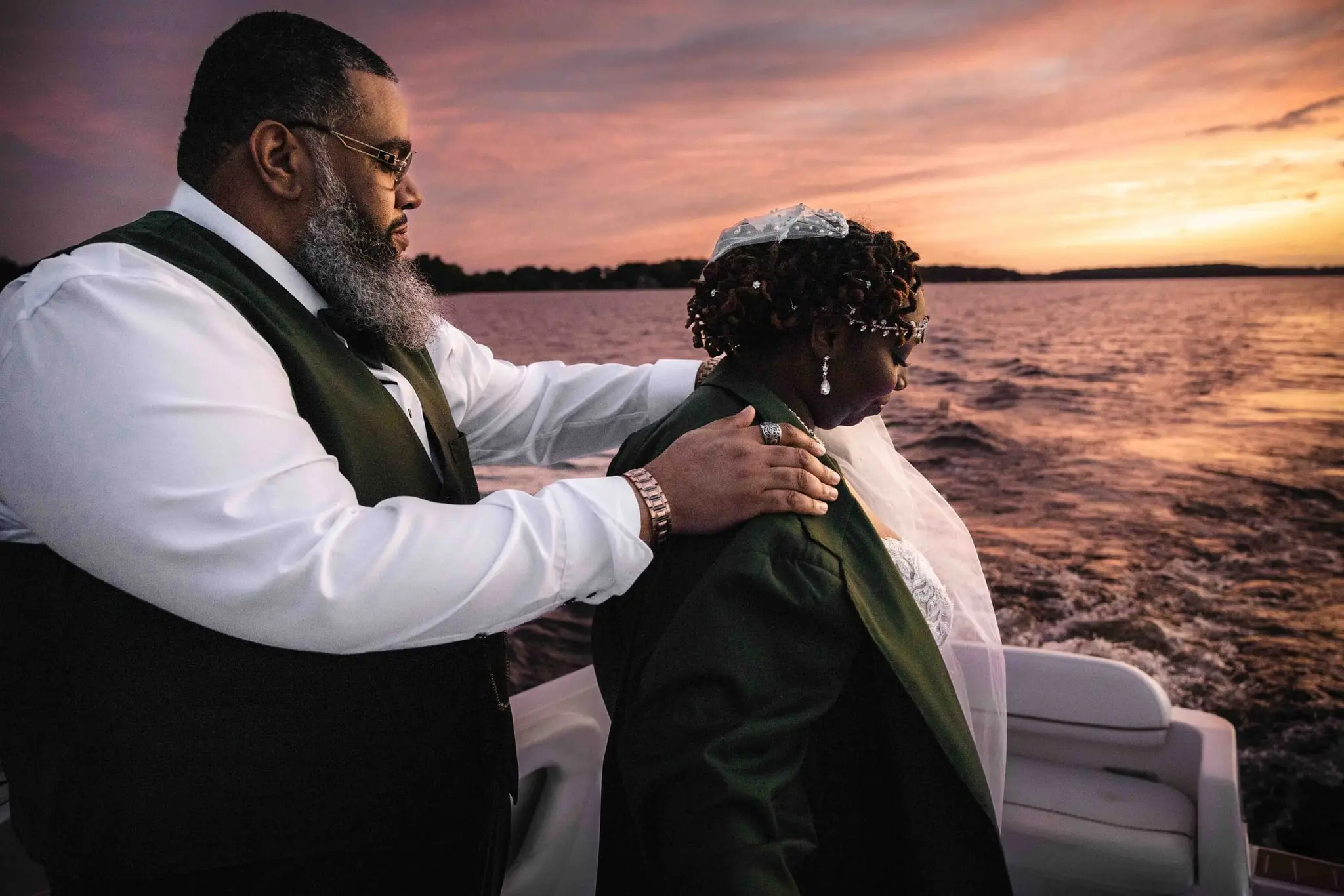 Groom settling his green jacket onto the bride's shoulders with the lake behind