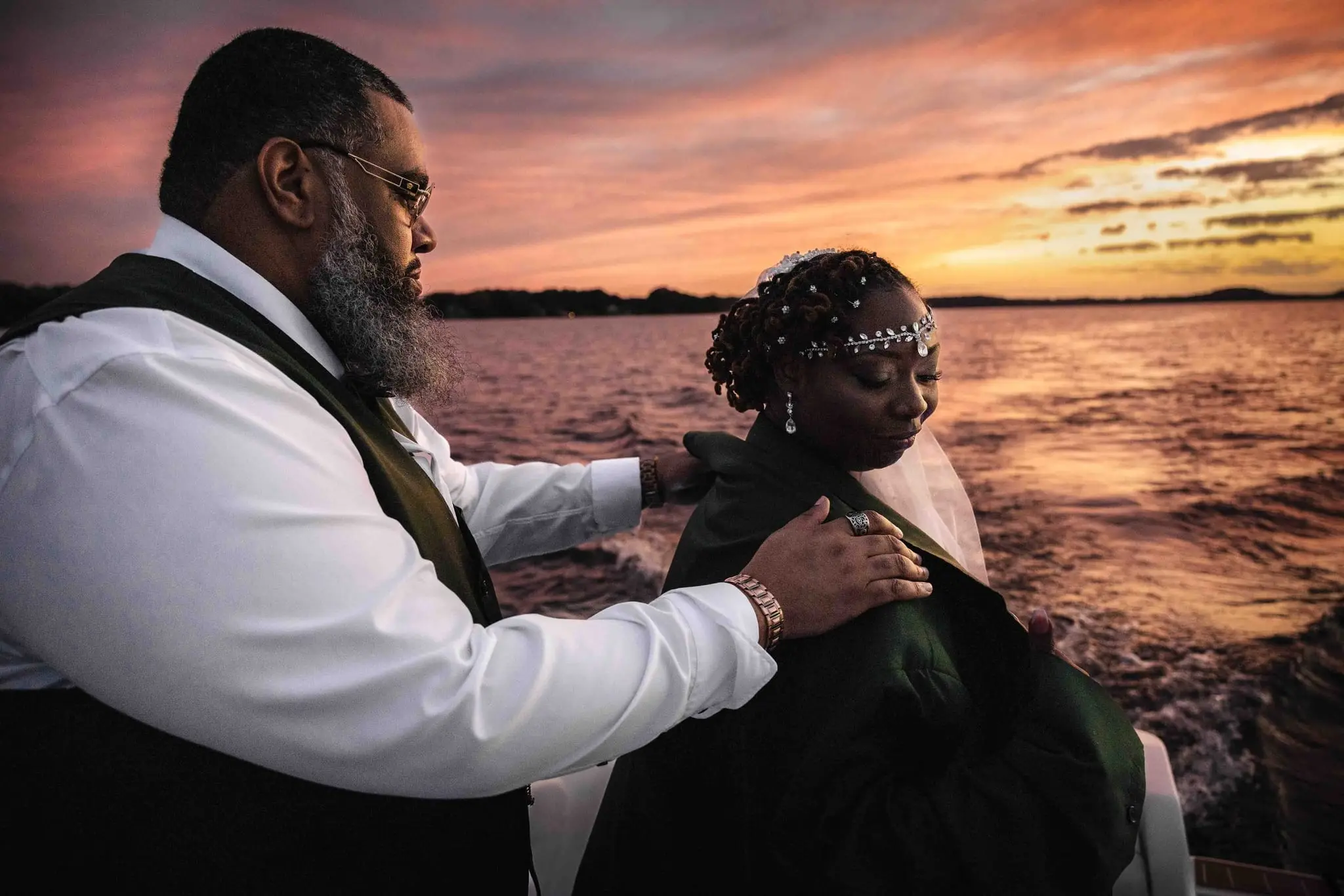 Groom draping his green jacket over the bride's shoulders on the lake at sunset