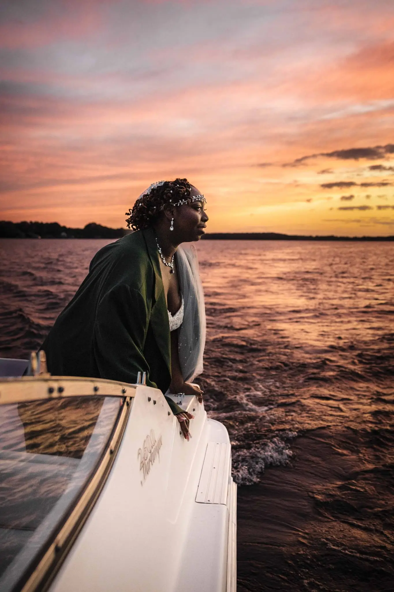 Bride leaning on the boat's side in the green jacket, watching the water and pink sunset