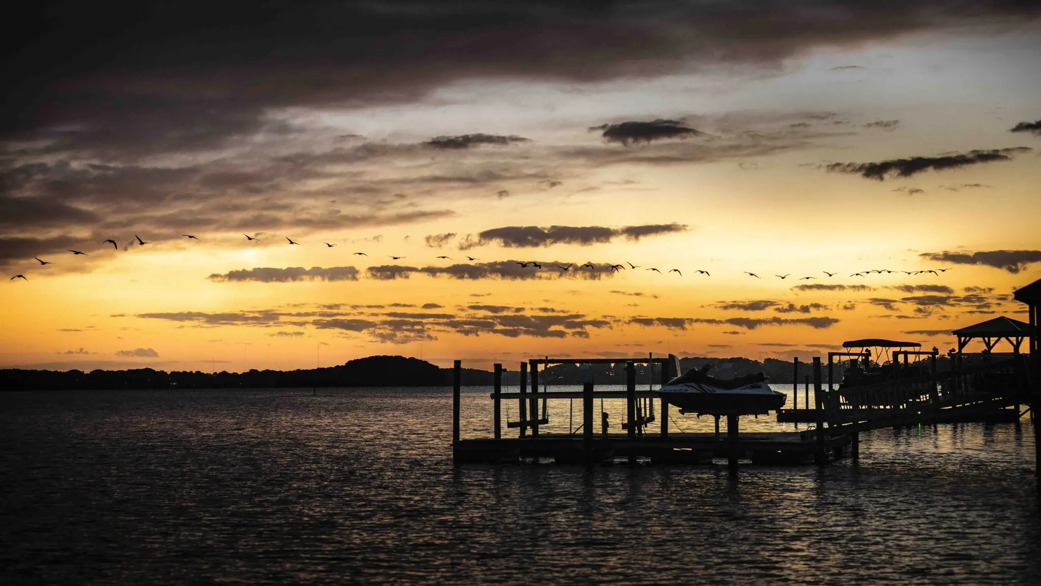 Geese flying across an orange sunset sky above a lakeside dock