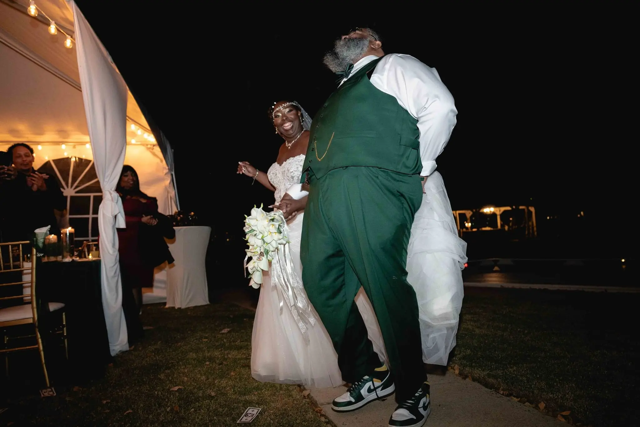 Groom dancing low in a green vest and sneakers as the bride laughs beside the reception tent