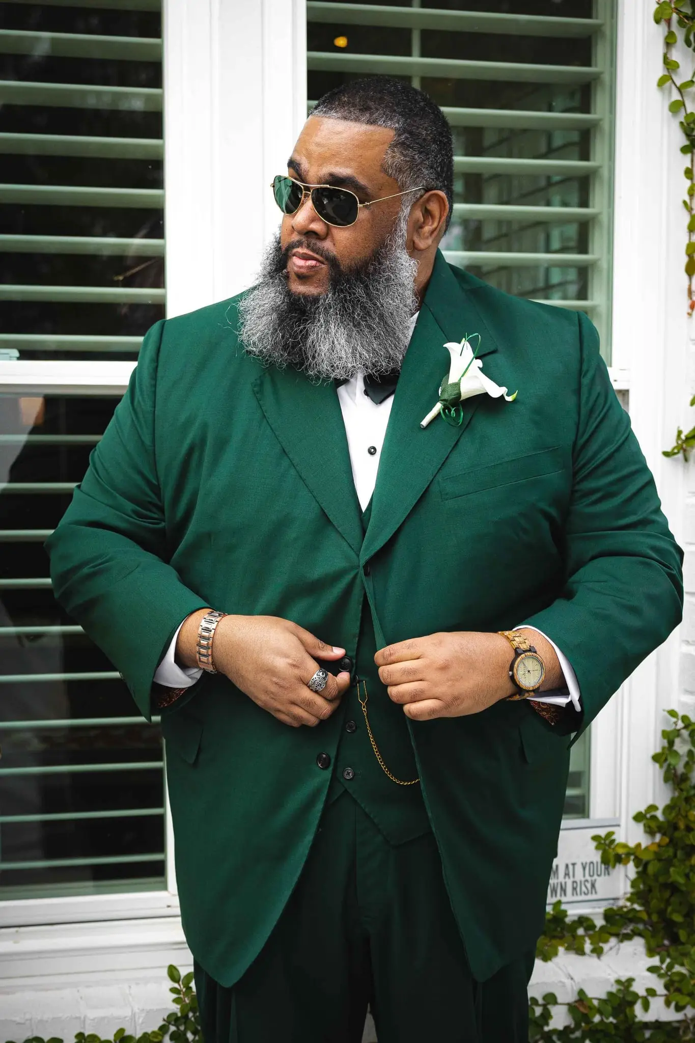 Groom in a green three-piece suit buttoning his jacket in front of green shutters
