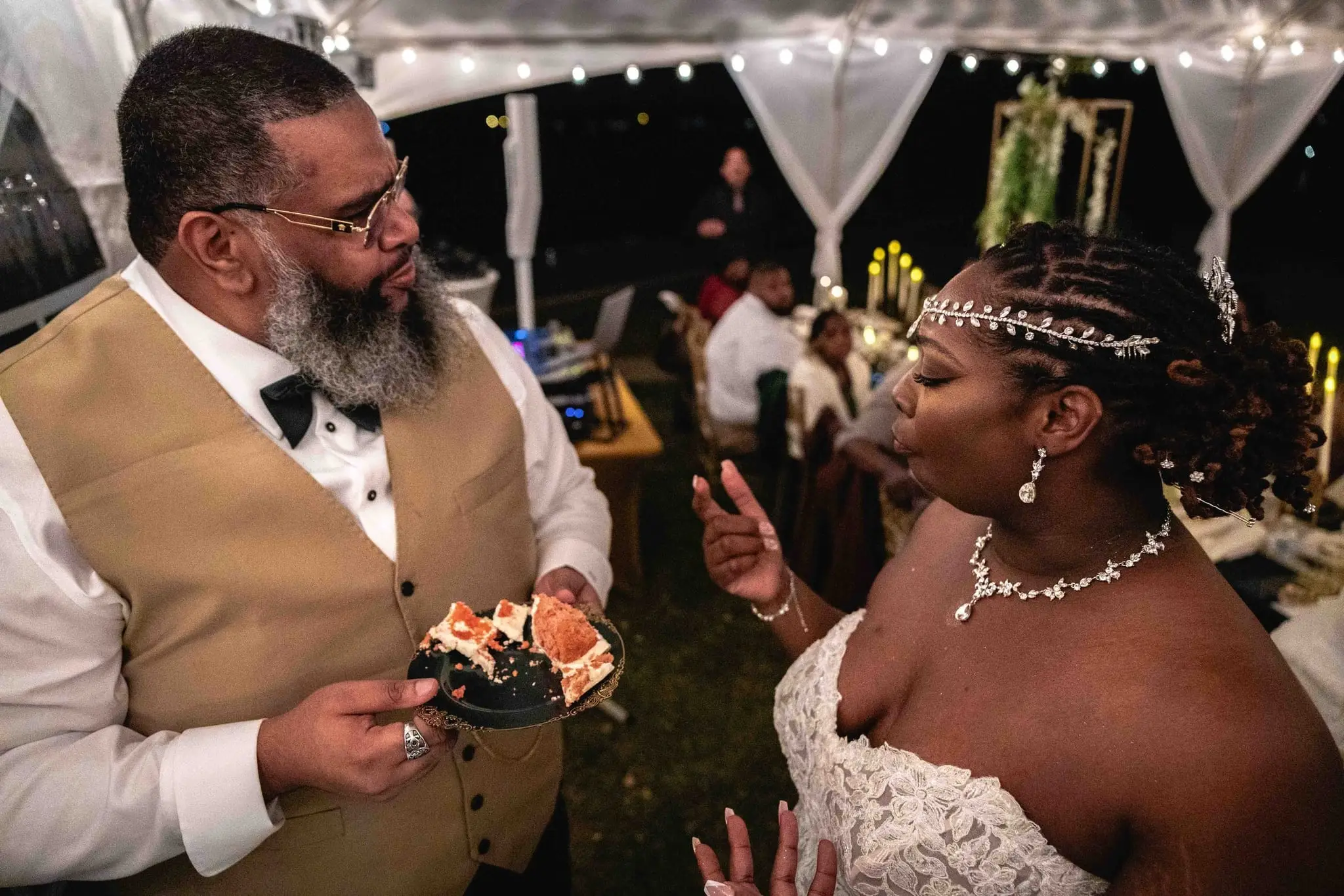 Groom in a gold vest holding a plate of cake while chatting with the bride under the tent