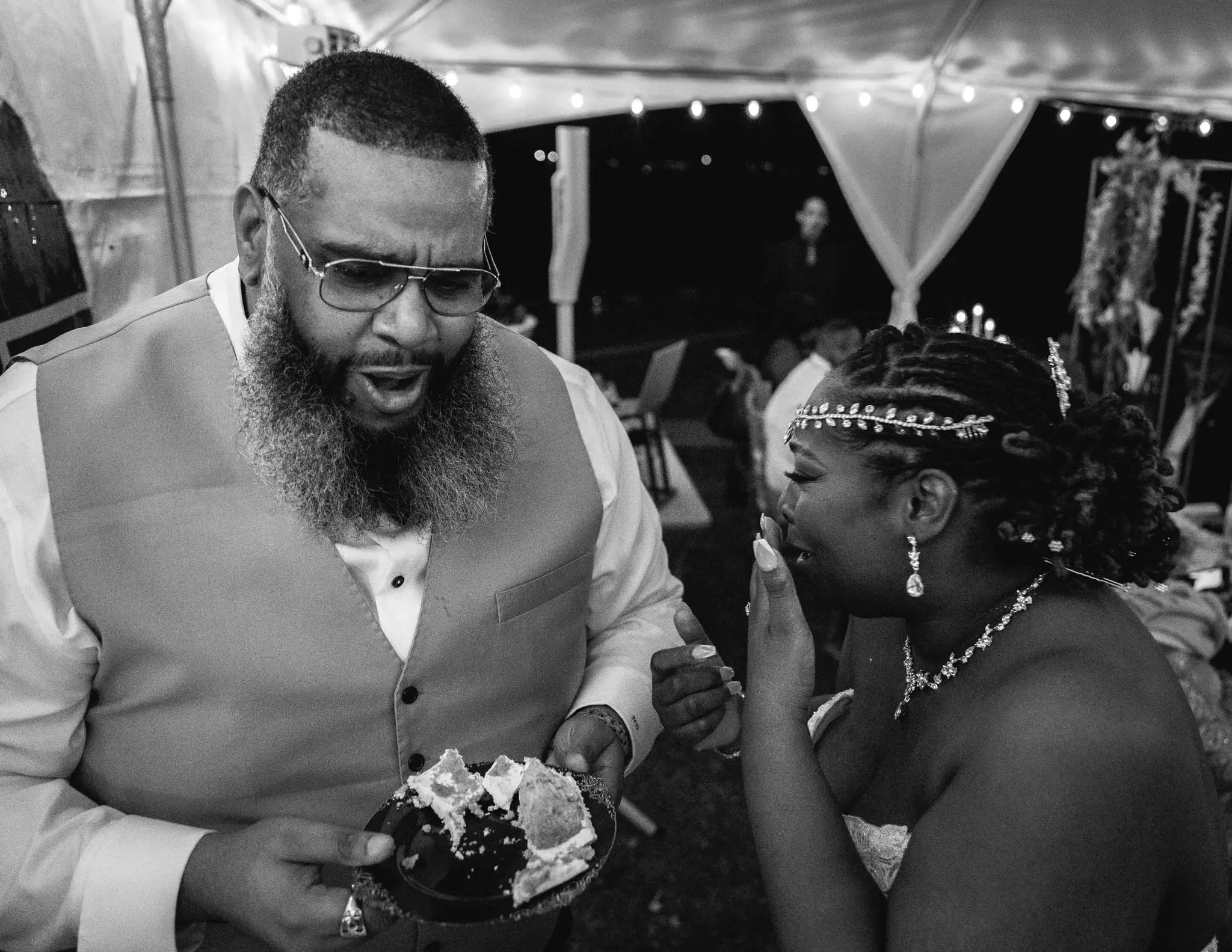 Black and white of the groom mid-sentence holding a cake plate as the bride covers her laugh