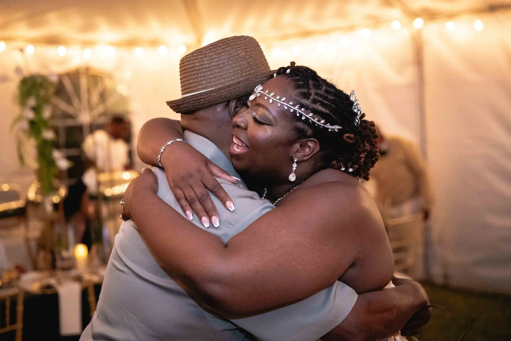 Bride laughing in a warm embrace with a guest in a fedora inside the lit reception tent