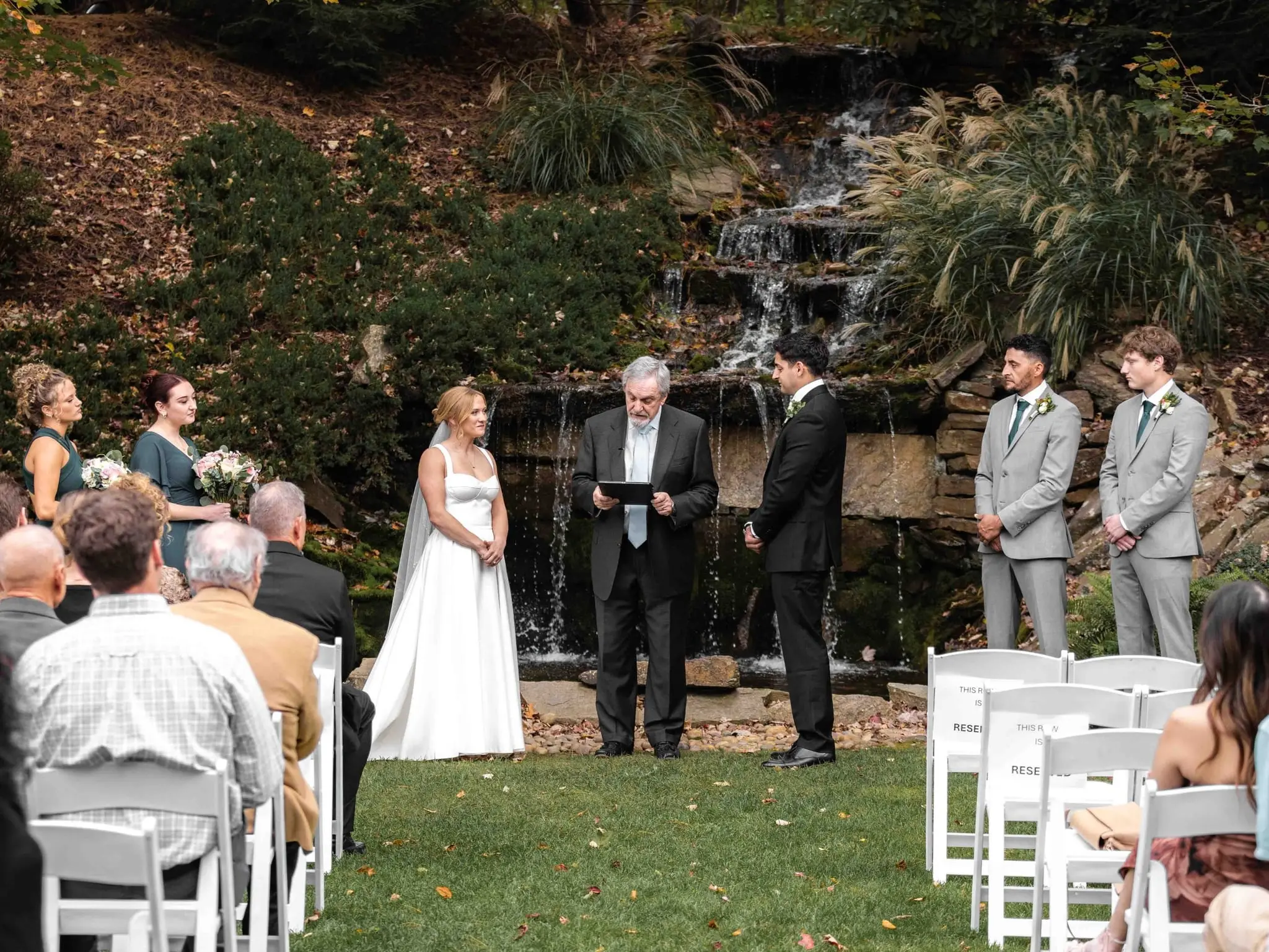 Lauren and Damian wedding at The Lodge at Flat Rock, Flat Rock NC — the ceremony on a lawn beside a small waterfall, the couple facing the officiant with guests on white chairs