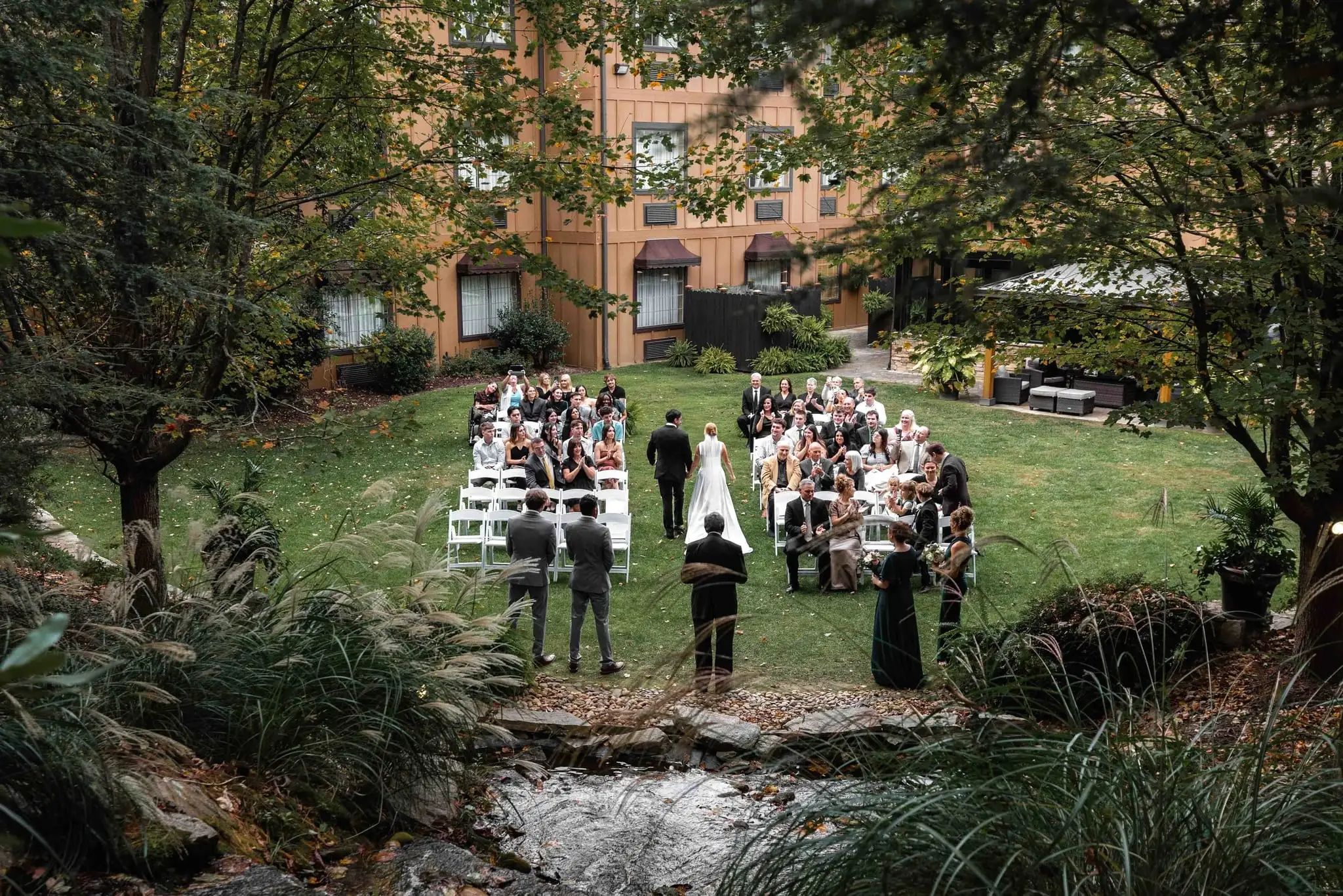 Wide view of a garden ceremony in the courtyard at The Lodge at Flat Rock, with a creek in the foreground