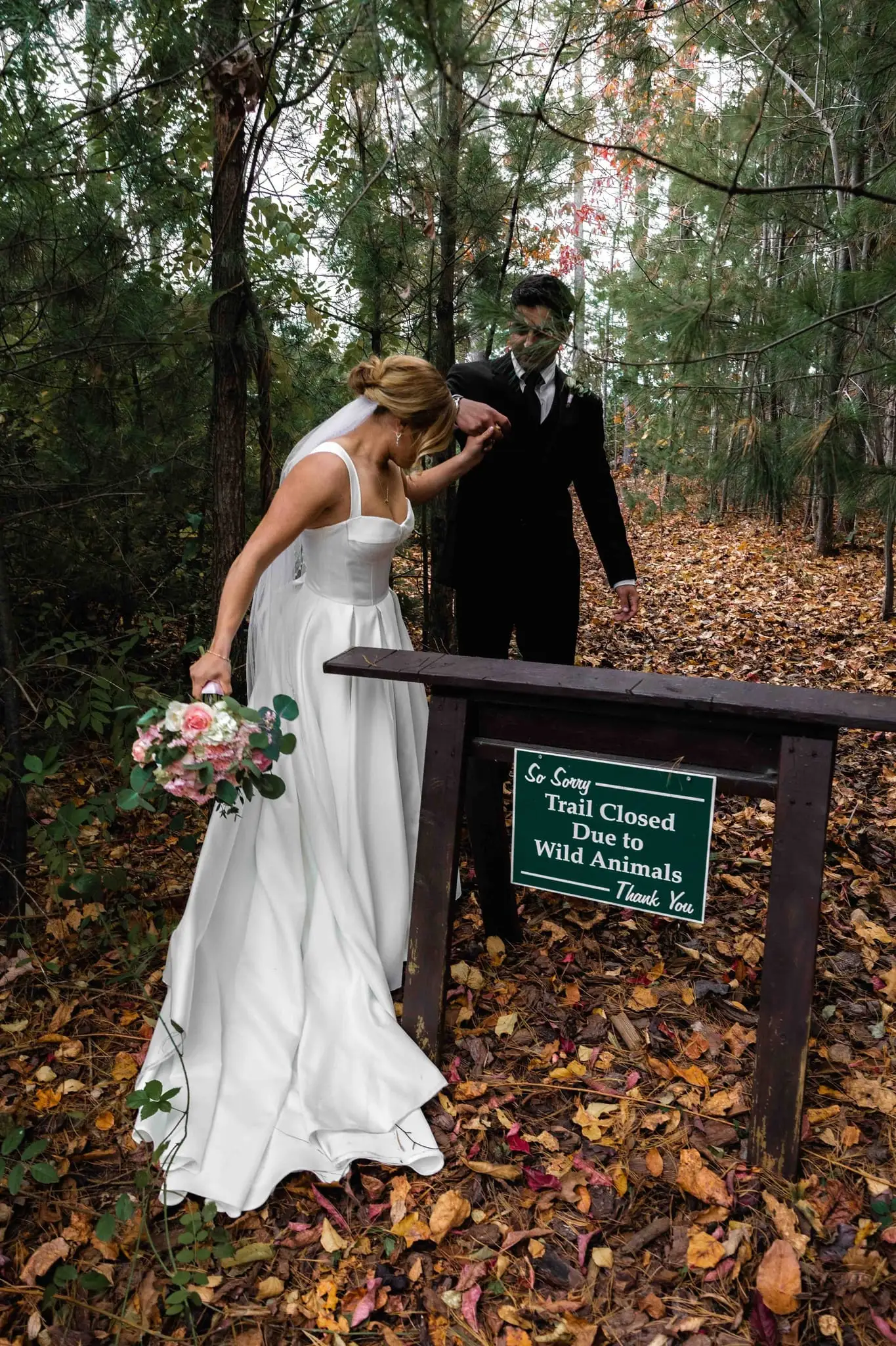 Lauren and Damian wedding at The Lodge at Flat Rock, Flat Rock NC — the bride bending to read a small green trail-closed sign in the leaves, bouquet in hand
