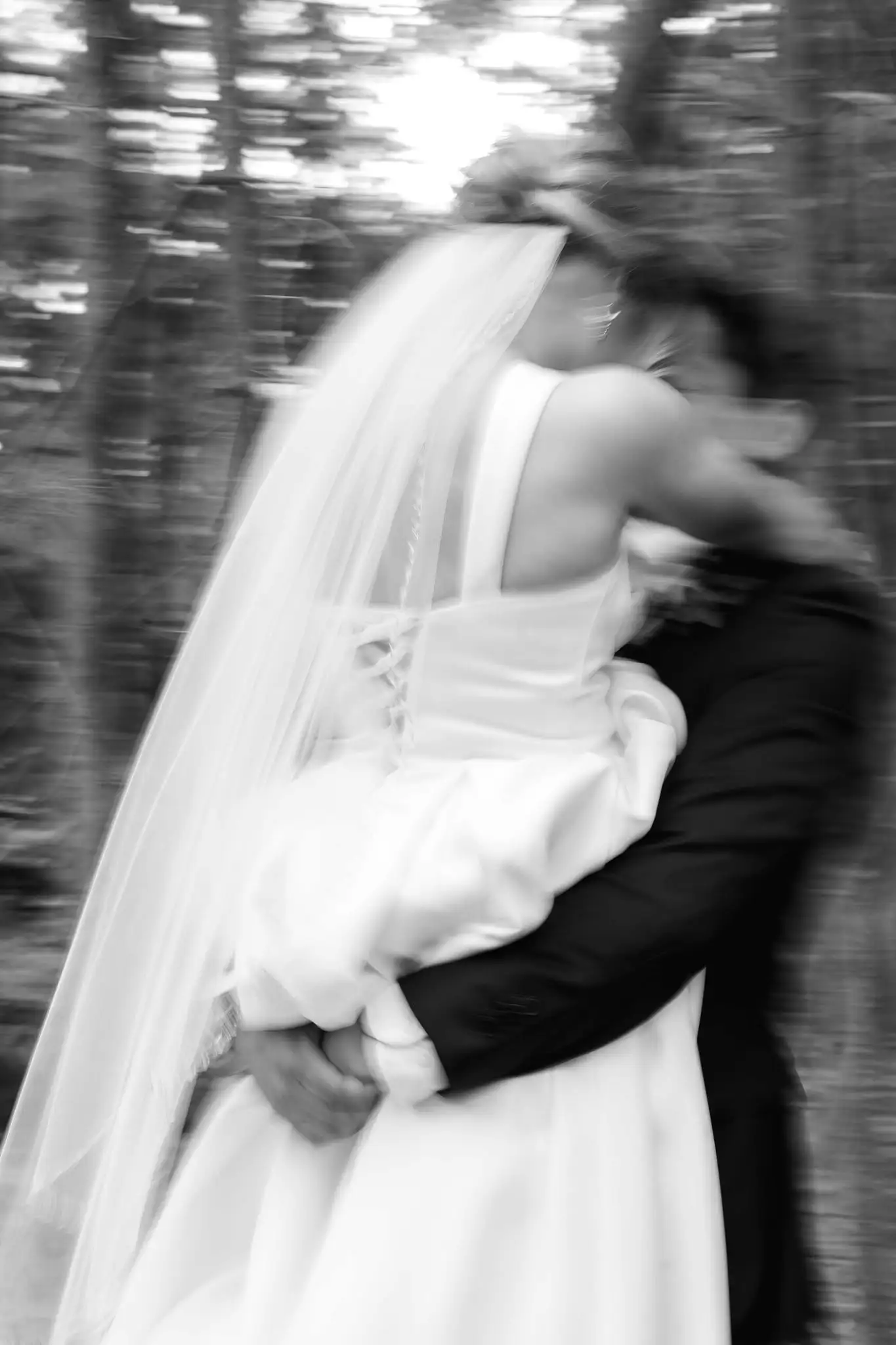 Black and white motion blur of the groom carrying the bride through the woods, her veil trailing