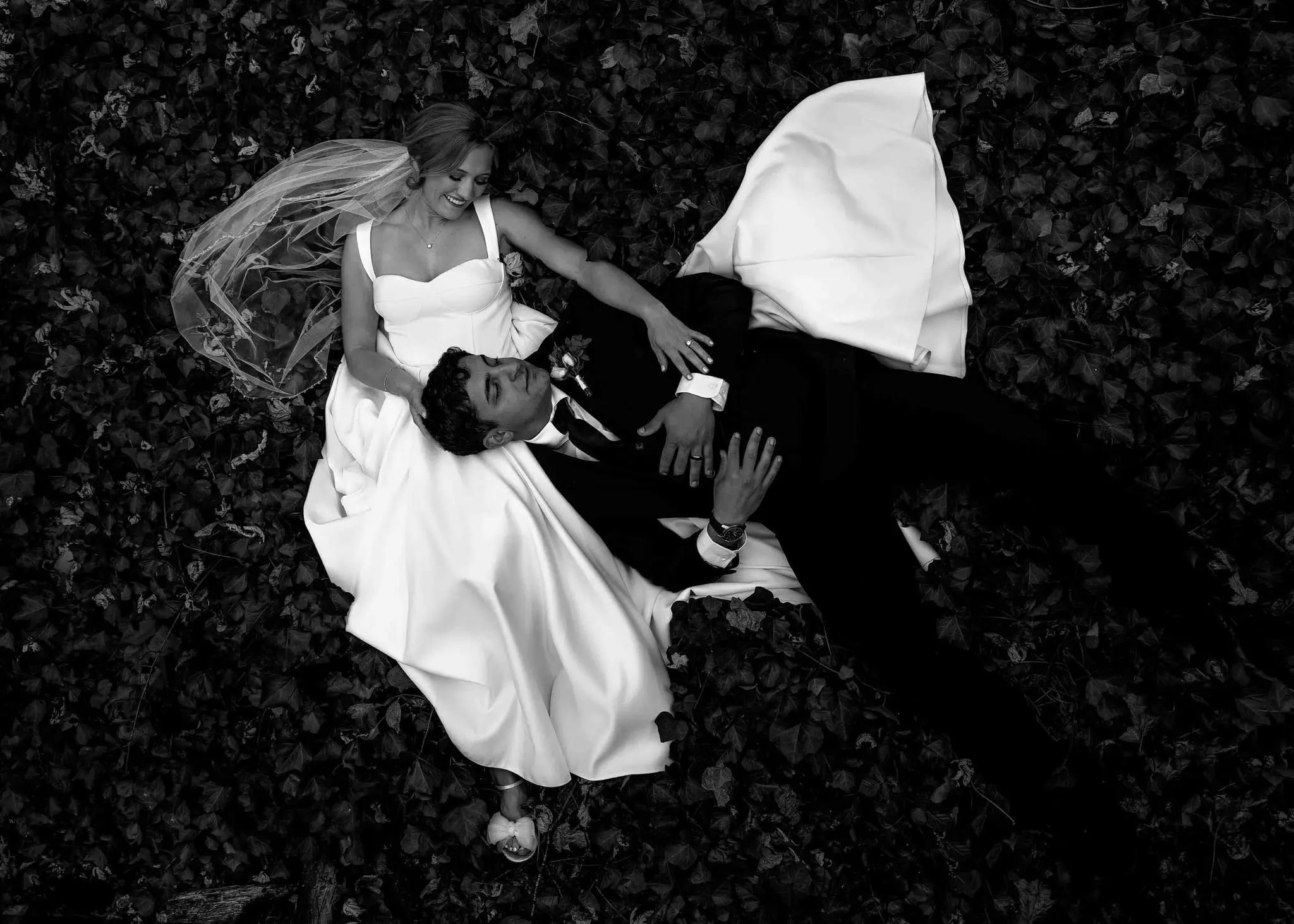 Black and white overhead shot of a bride and groom lying on fallen leaves, her veil spread beside them