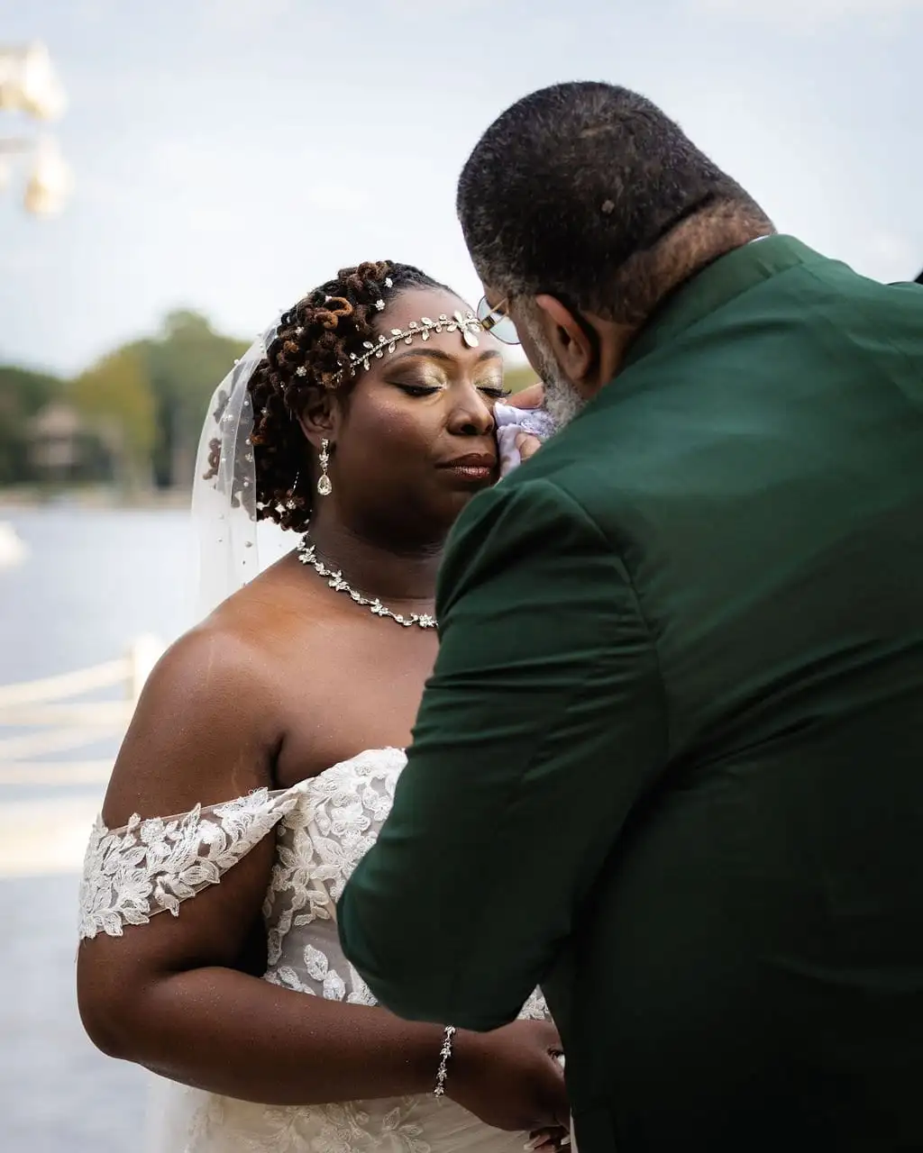 Bride in a beaded headpiece and groom in a green jacket share a quiet moment beside a lake