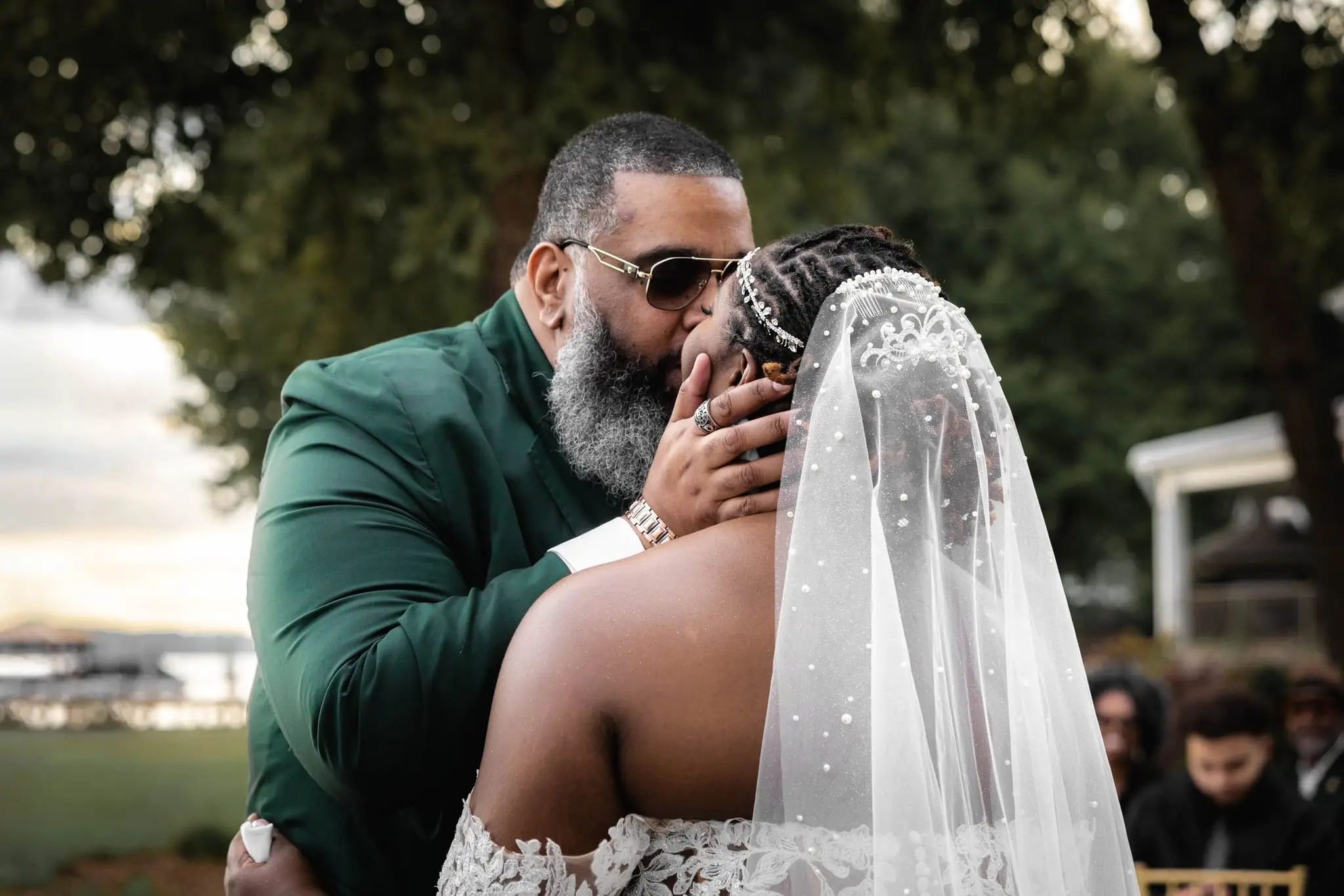 Groom in a green suit and sunglasses kissing the bride under her pearl-studded veil at an outdoor ceremony