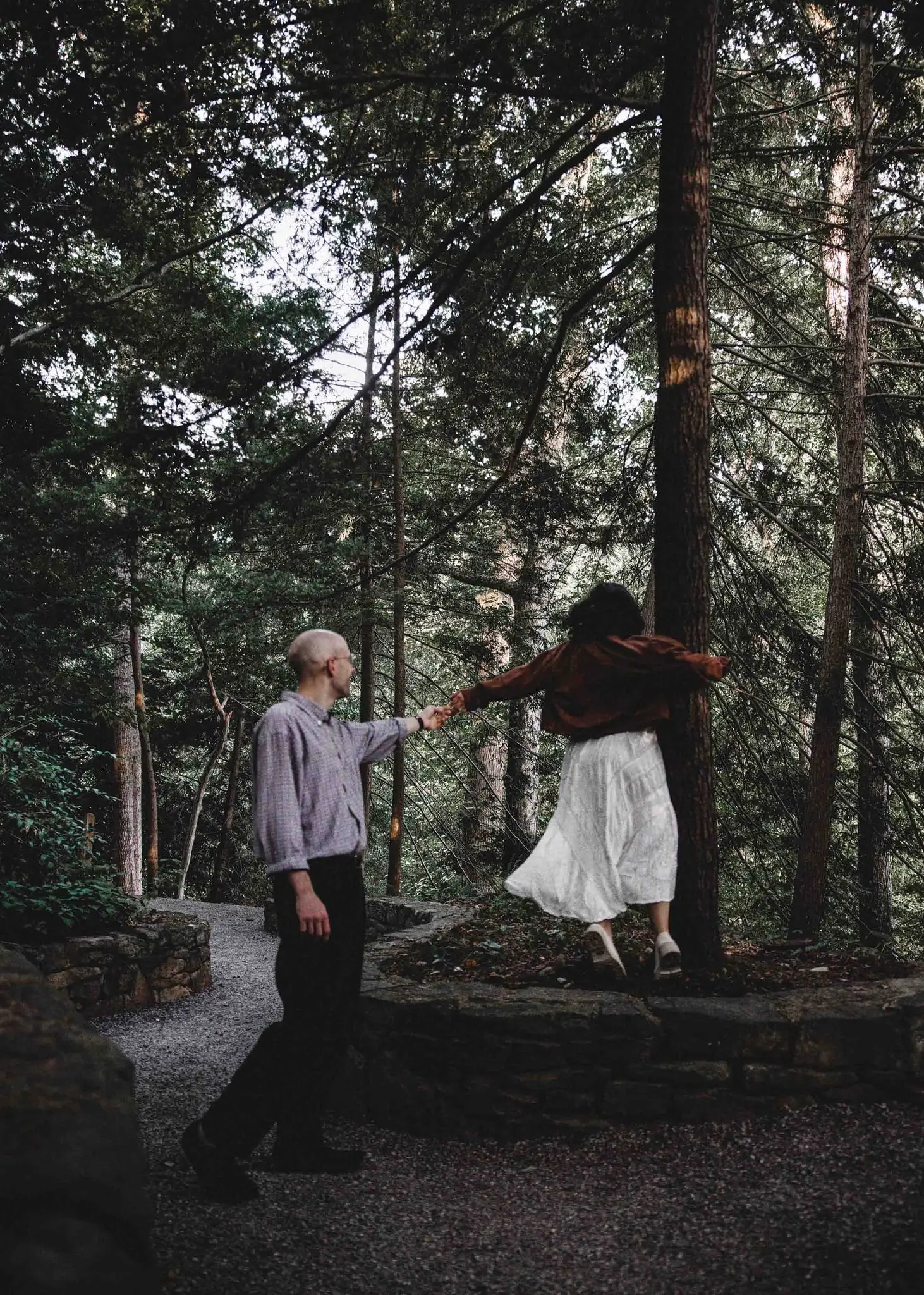 Man in a gray shirt pulling a woman in a white skirt and brown jacket along a woodland path