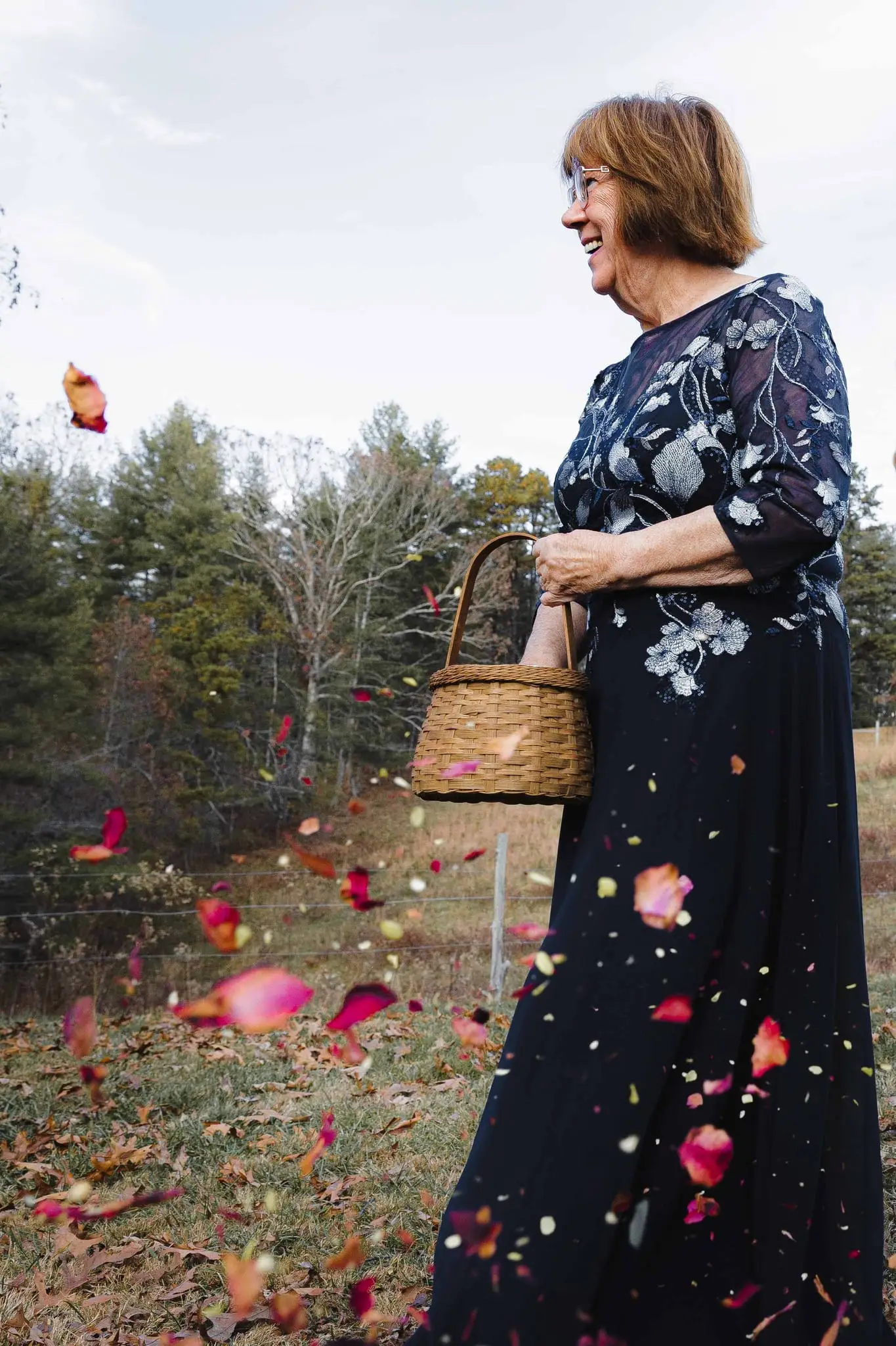 Smiling guest in a navy floral dress scatters rose petals from a basket on the lawn
