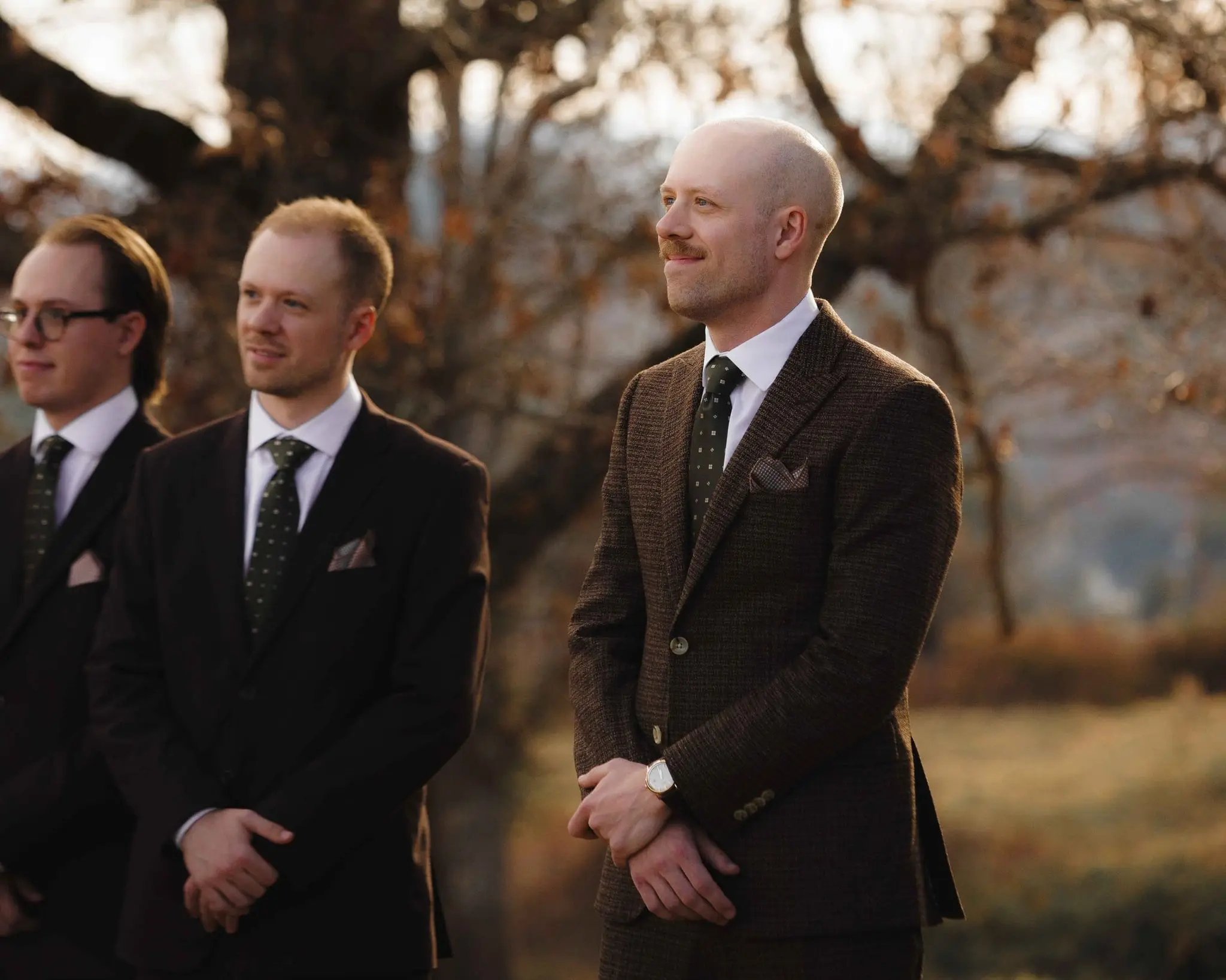 Groom in tweed grins at the end of the groomsmen line at the ceremony site in golden light