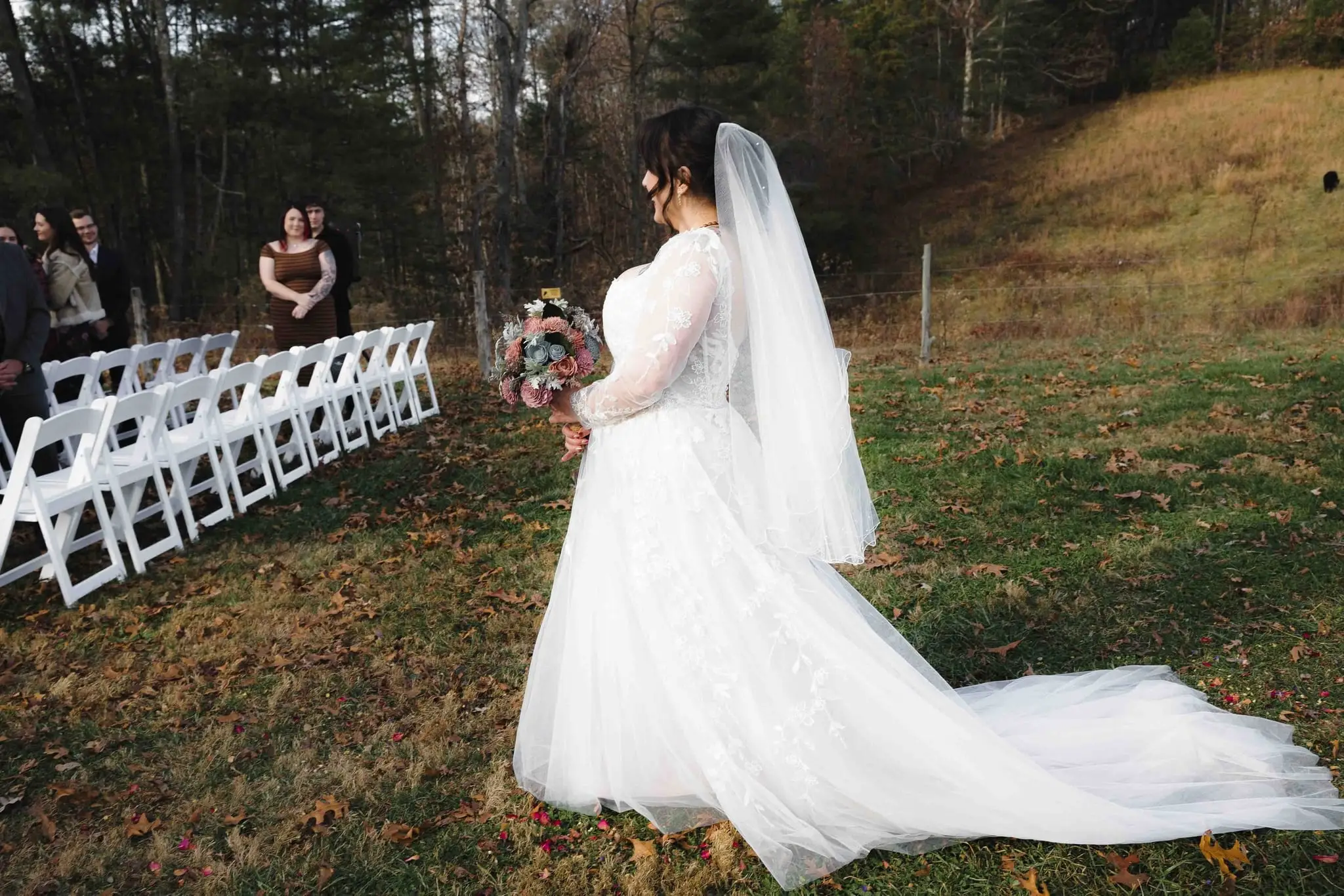 Elizabeth and Aidan wedding at Emerald Ridge Farm, Weaverville NC — the bride seen from the side, her long veil trailing, facing rows of white folding chairs
