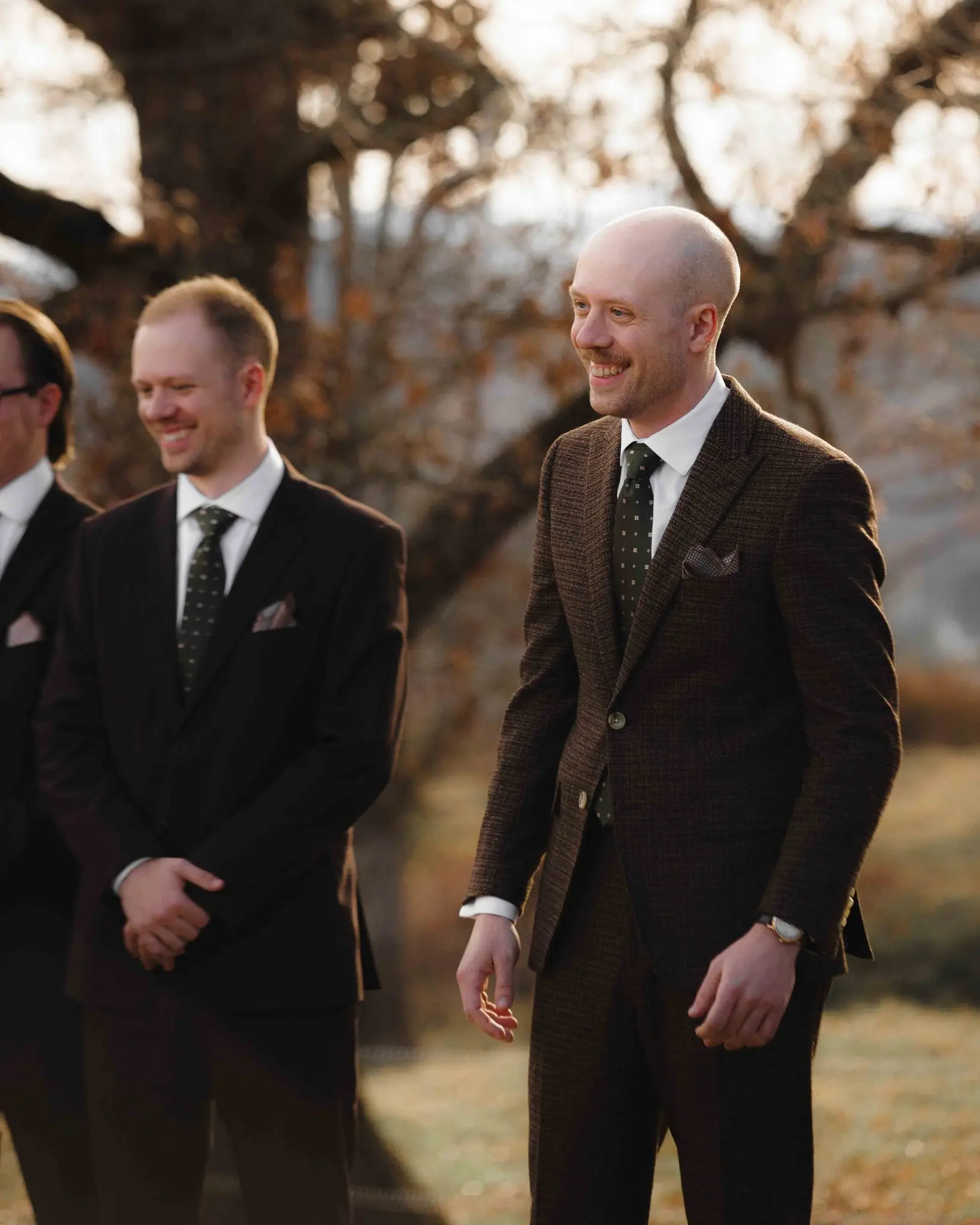 Groom laughs with groomsmen beside him as he waits at the ceremony under bare oak trees