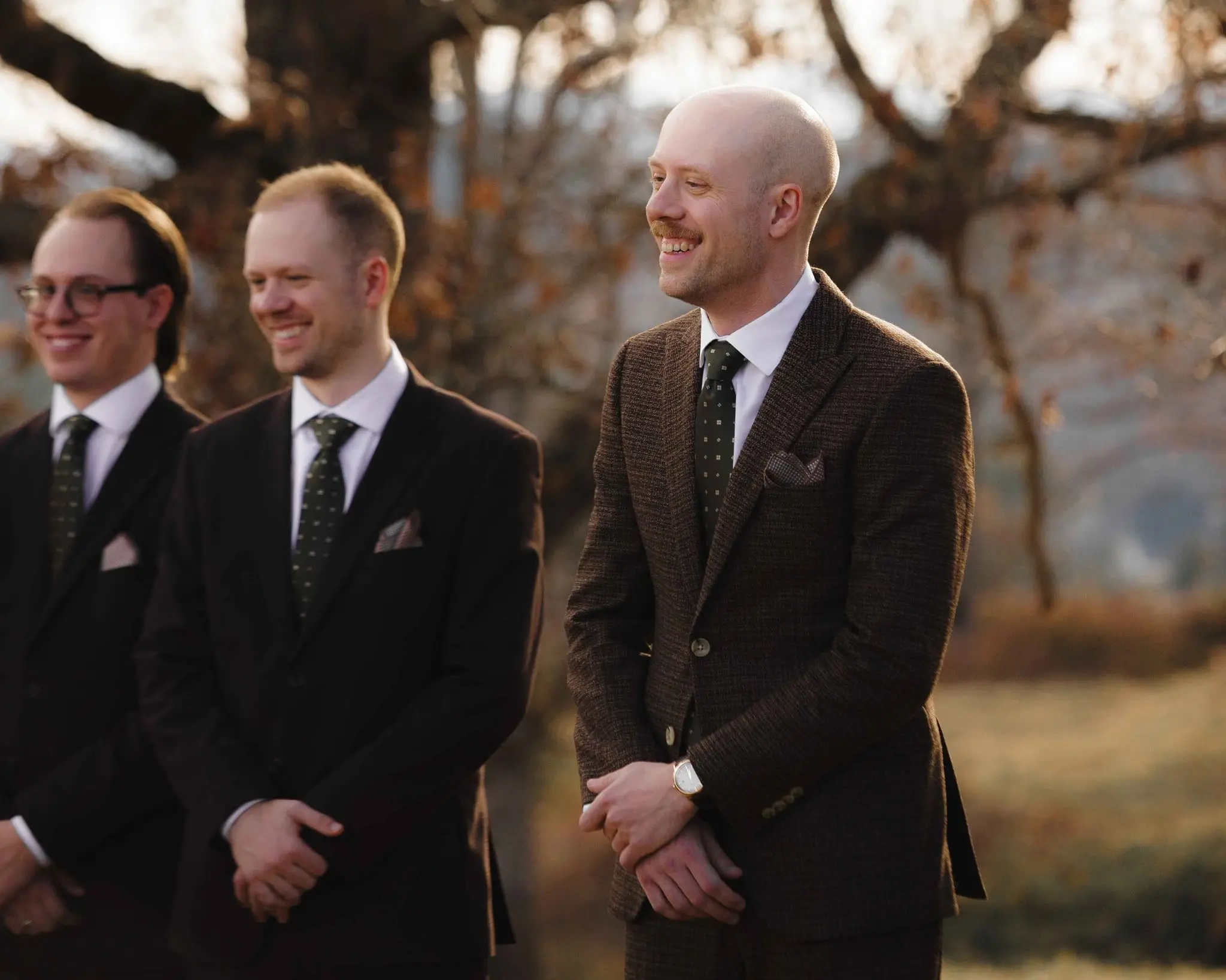 Groomsmen smile as they watch the bride walk the aisle, the groom in a brown tweed suit at right