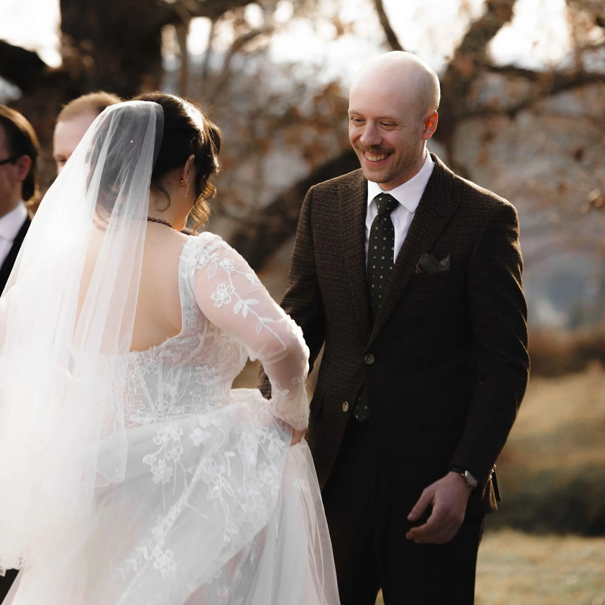 Groom grins as the bride arrives, her veil and lace sleeves catching the evening light
