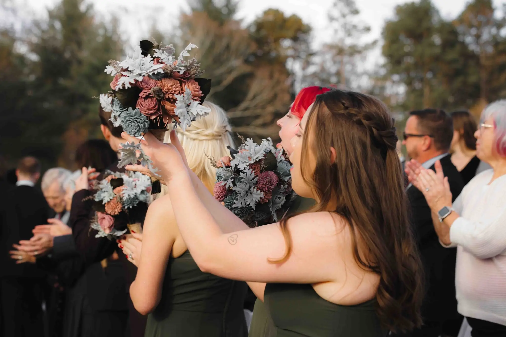 Guests clap while a bridesmaid in green holds a dusty rose bouquet high during the ceremony