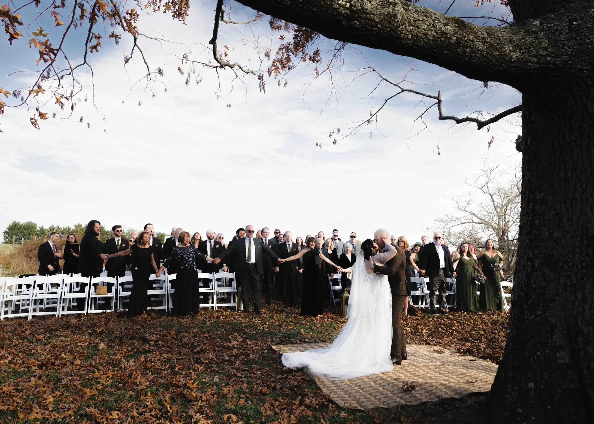 Elizabeth and Aidan wedding at Emerald Ridge Farm, Weaverville NC — the ceremony in a field, the couple at the front beneath a large bare tree with guests seated on white chairs