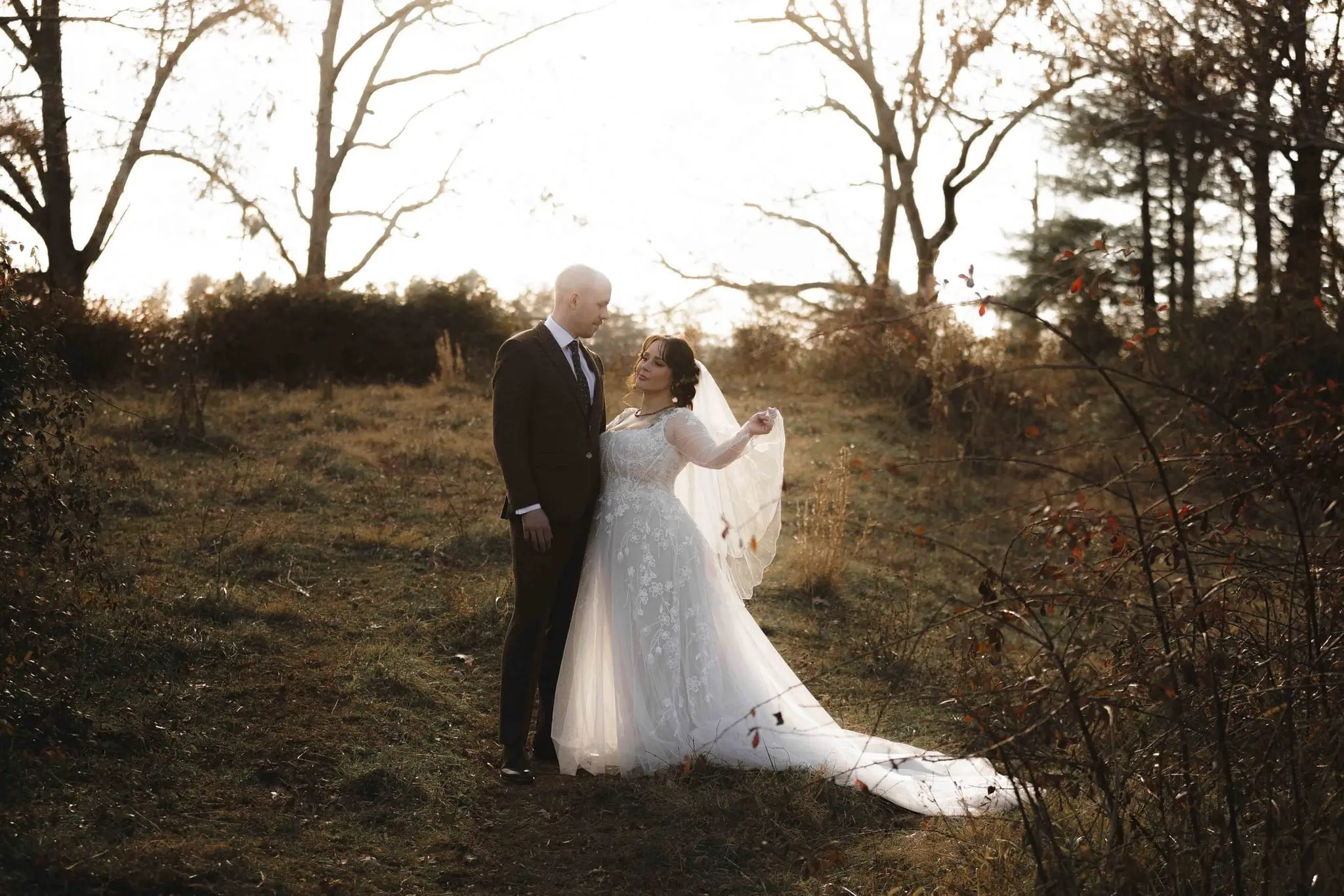 Couple stand face to face in a field at golden hour, the bride lifting the edge of her veil
