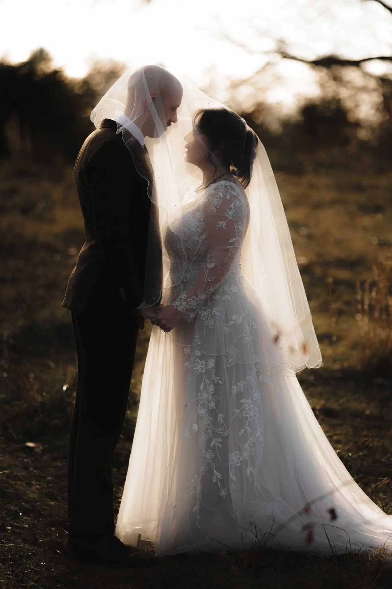 Bride and groom hold hands in the warm glow of sunset beneath the bride's long veil