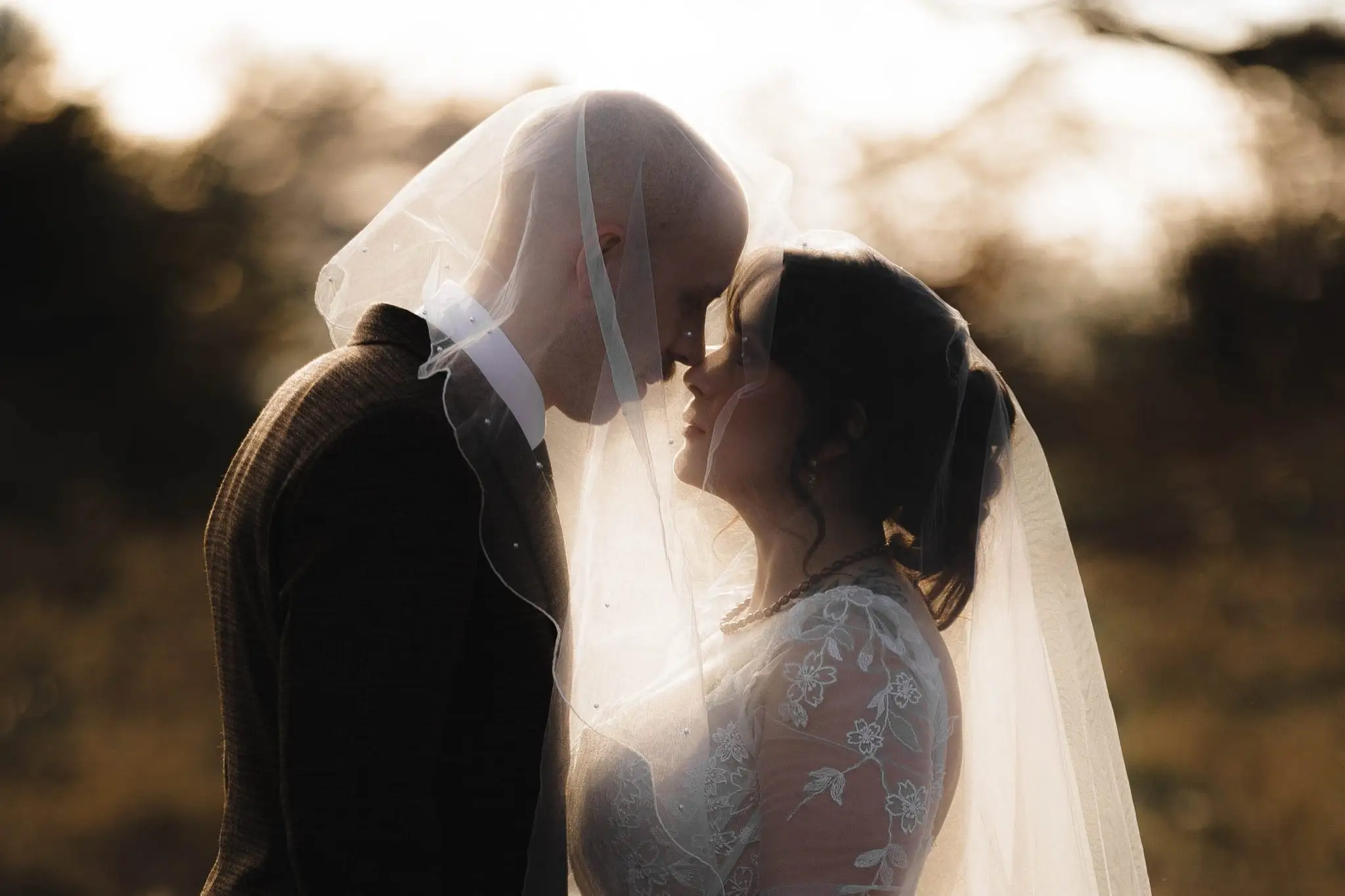A couple touching foreheads under a veil, backlit by low golden sun