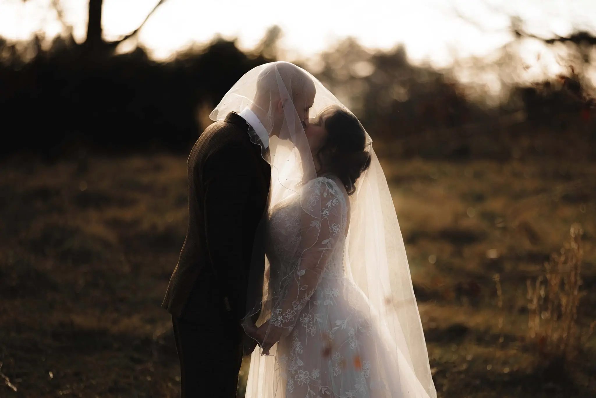 Couple kiss beneath the sheer veil in a field with golden light behind them
