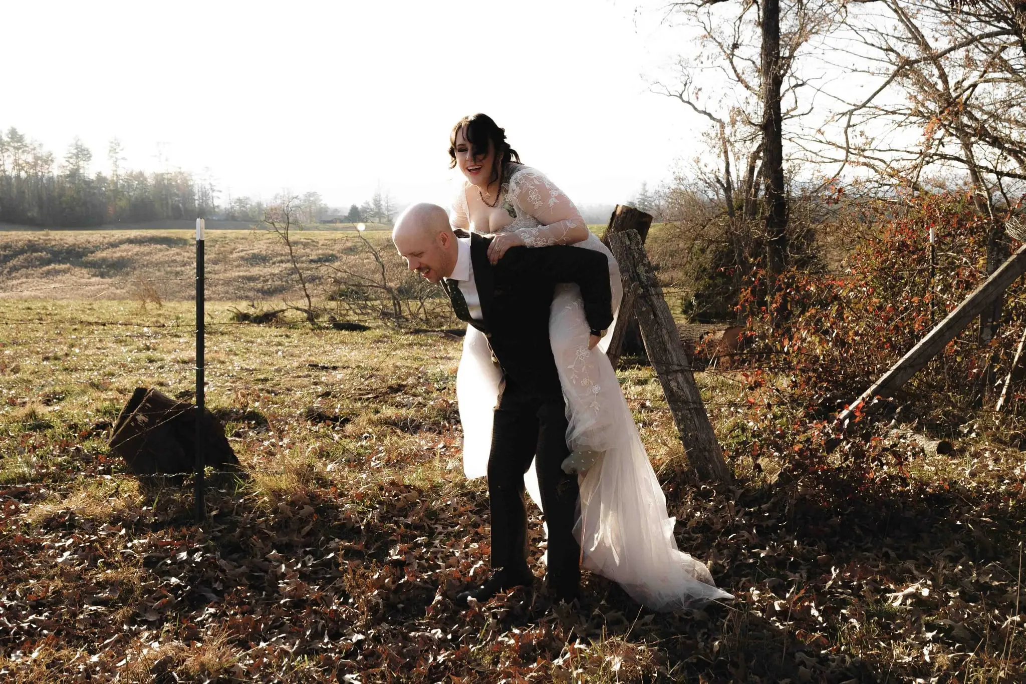 Groom bends forward carrying the bride piggyback across a fenced pasture