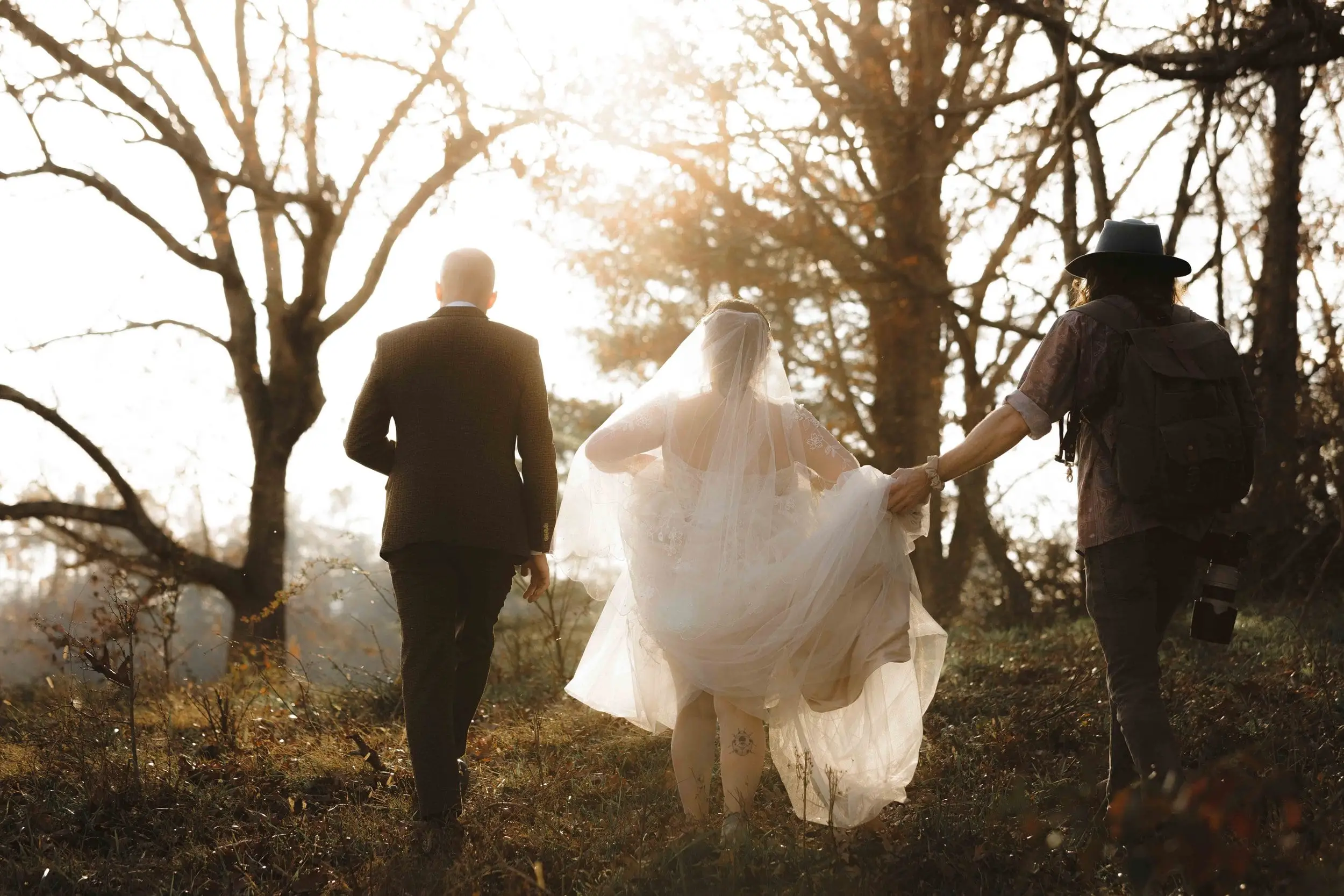 A bride and groom walking away through bare trees toward the late-afternoon sun with a guest beside them