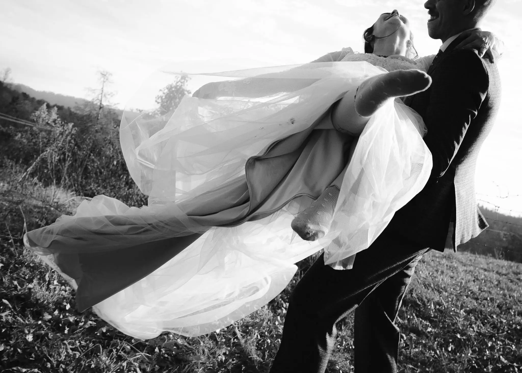 Black and white shot of the groom carrying the bride in his arms, her dress billowing in the wind