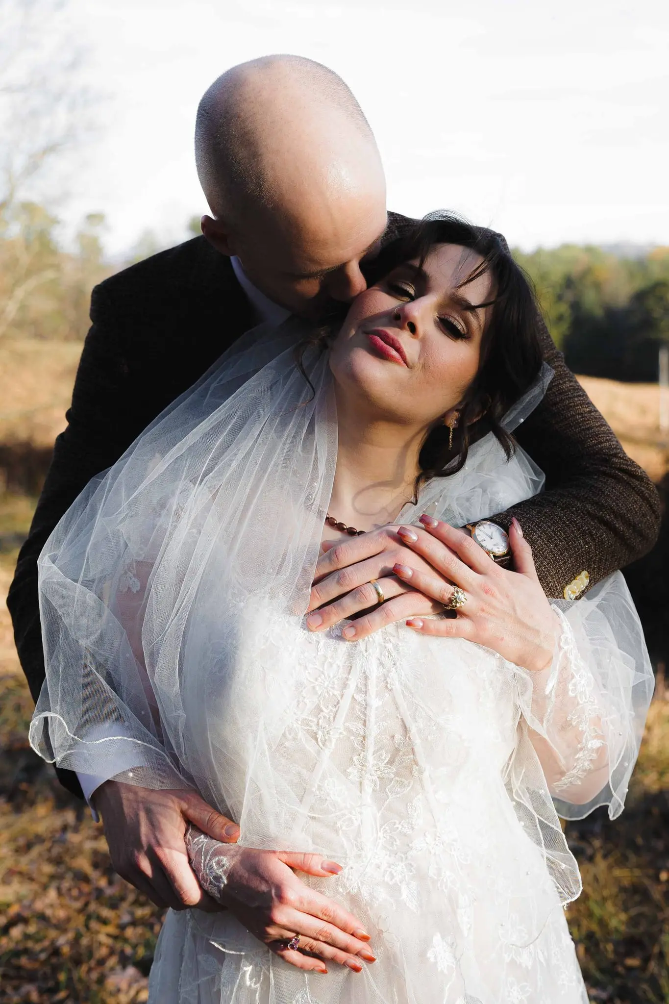 Groom kisses the bride's cheek from behind as she smiles with her hands on her heart
