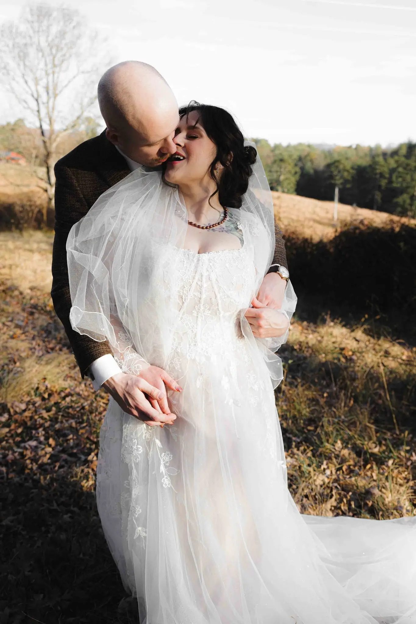 Groom nuzzles the bride's neck as she laughs, her veil gathered around her arms