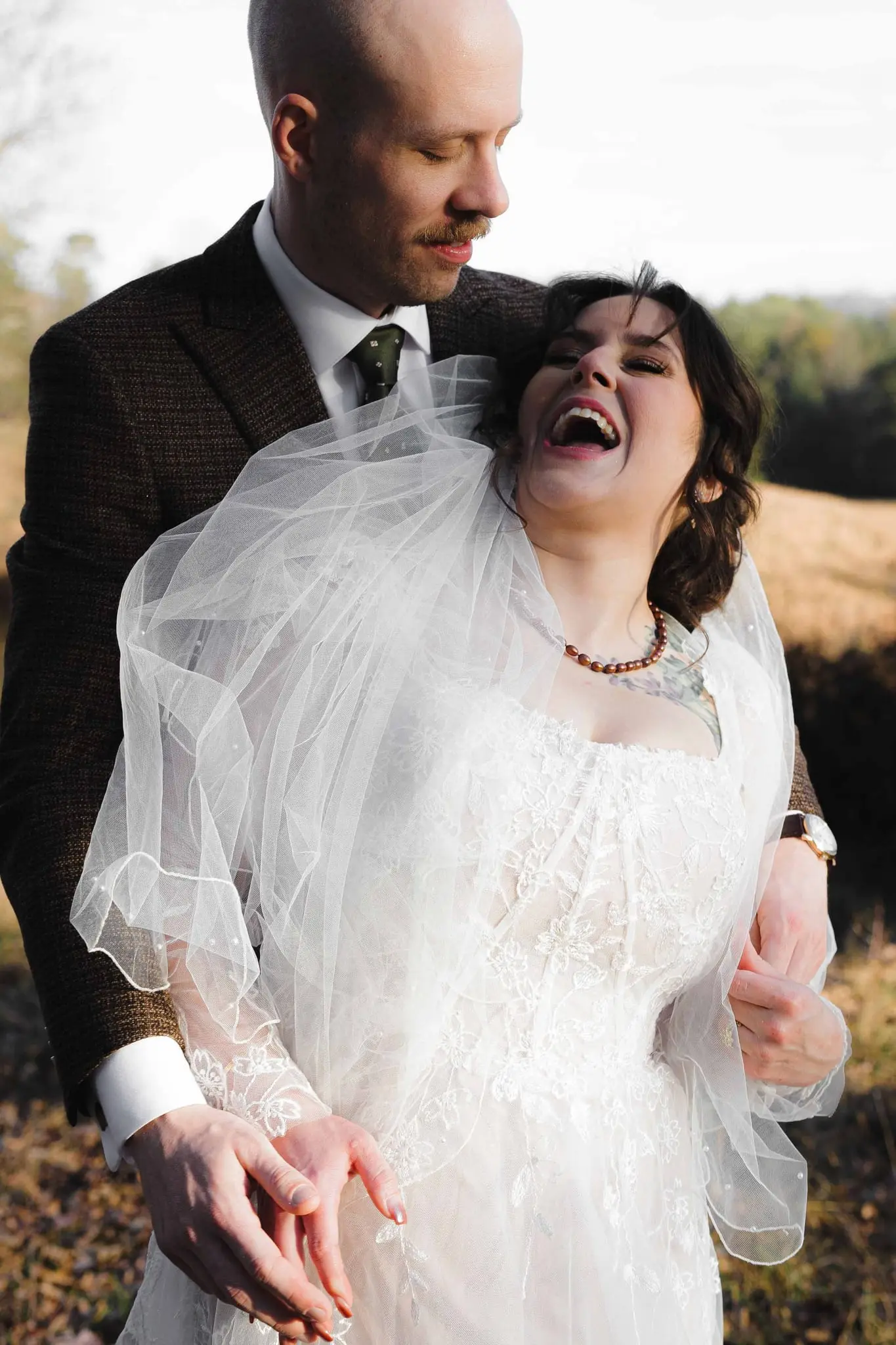 Groom hugs the bride from behind as she laughs, her veil gathered around them in an autumn field