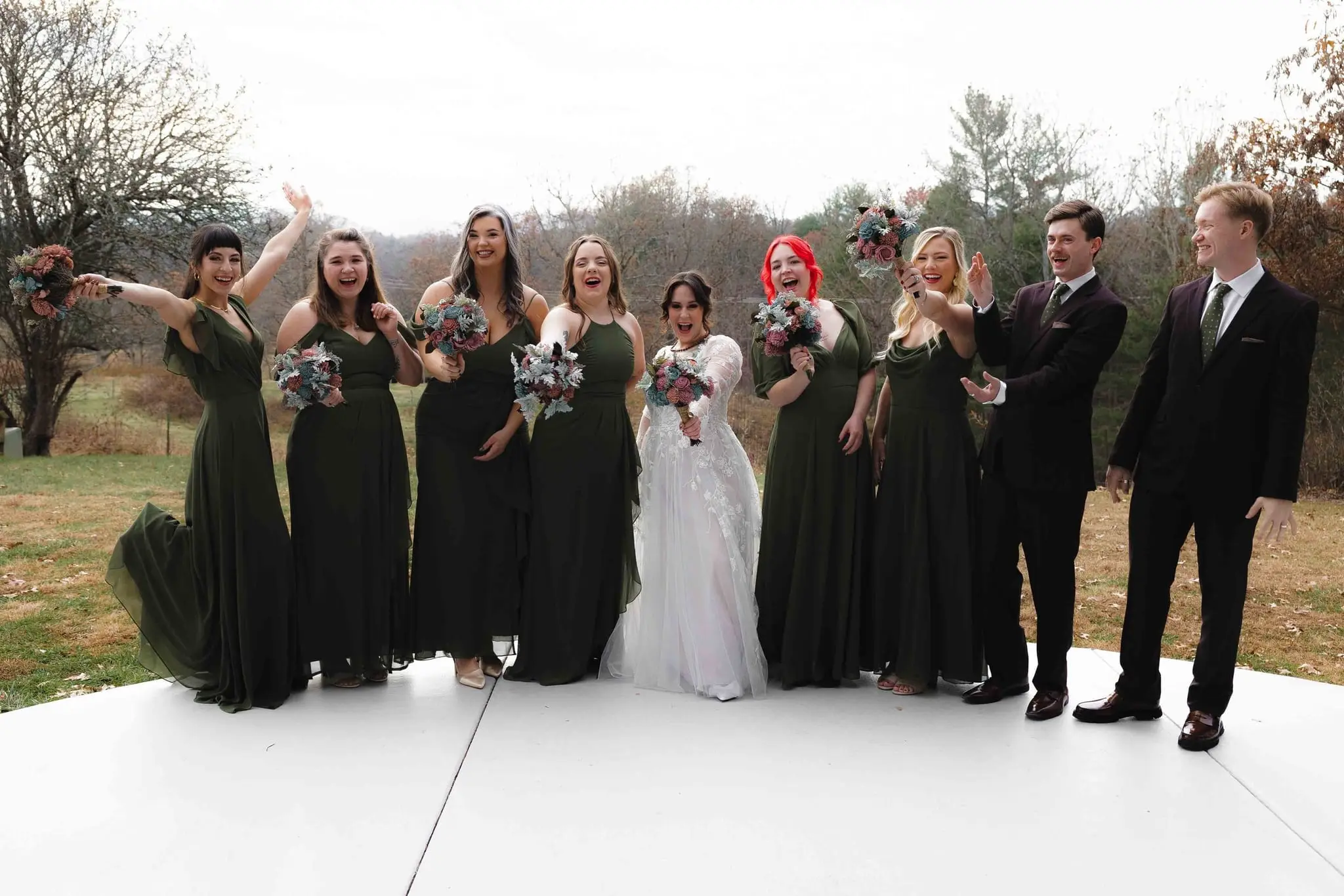 Bridal party in forest green cheering and tossing their arms up on a white platform